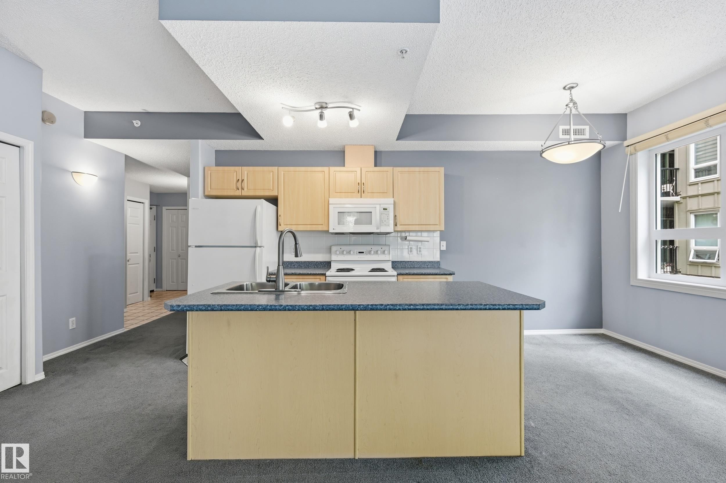 Open concept kitchen featuring an island with an integrated double basin sink, light wood-finish cabinetry, and white appliances - 203 11103 84 Avenue, Edmonton, AB
