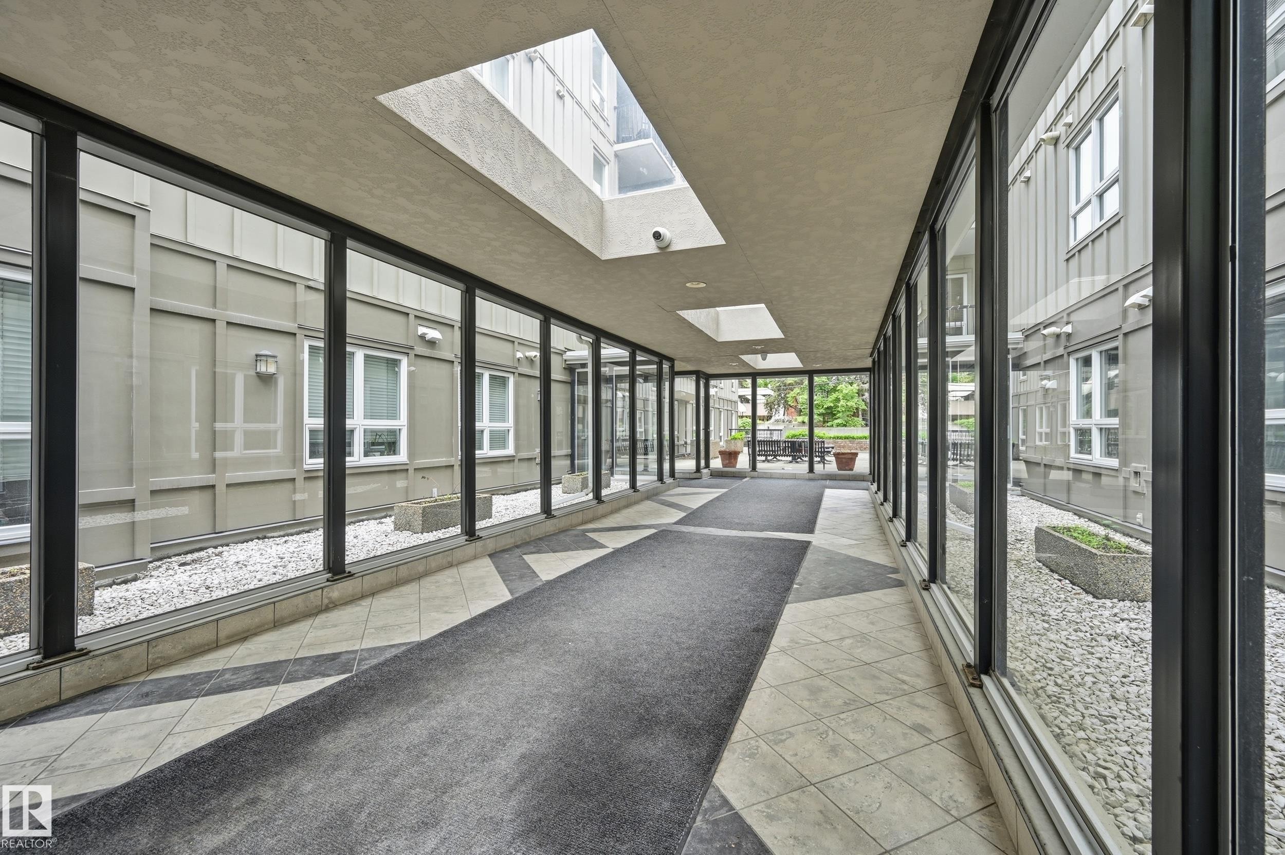 Enclosed walkway featuring a patterned tile floor with a carpet runner, multiple skylights, and full-height windows on both sides - 203 11103 84 Avenue, Edmonton, AB