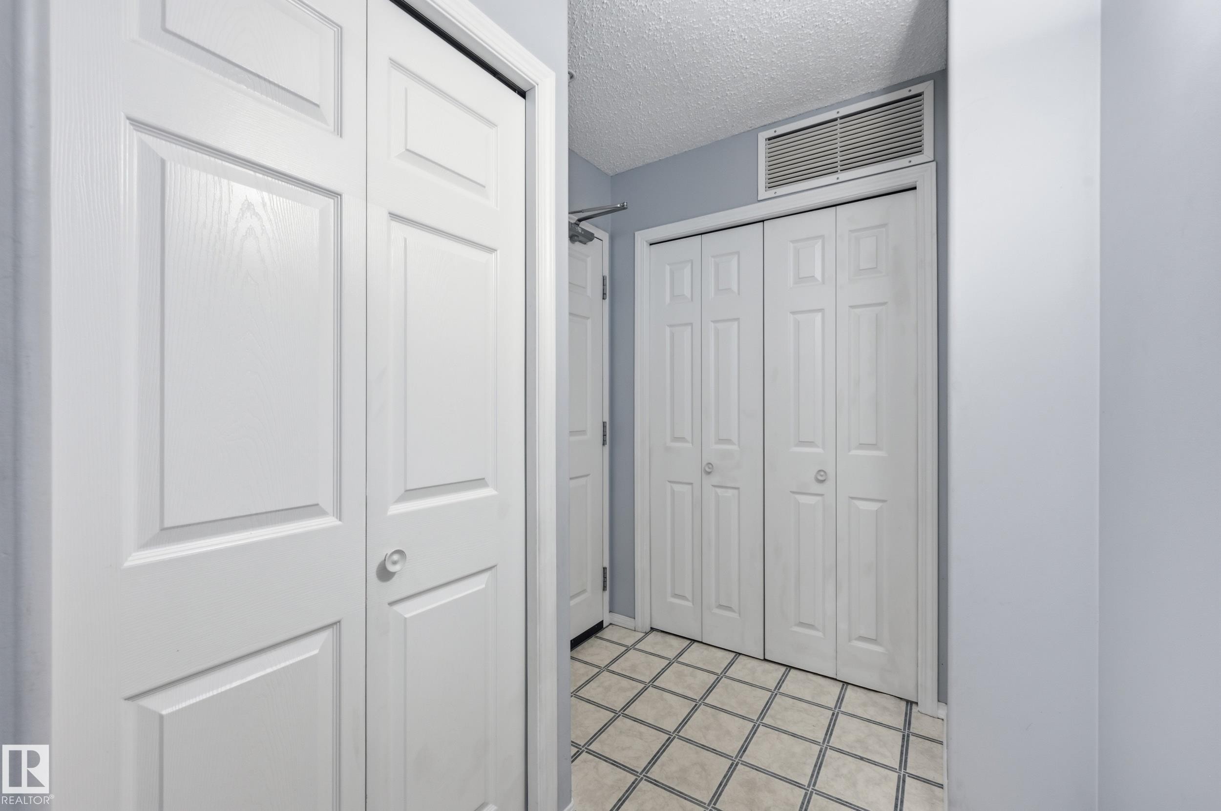 Hallway featuring panel-style bifold closet doors, light blue wall paint, and square tile flooring with contrasting grout - 203 11103 84 Avenue, Edmonton, AB