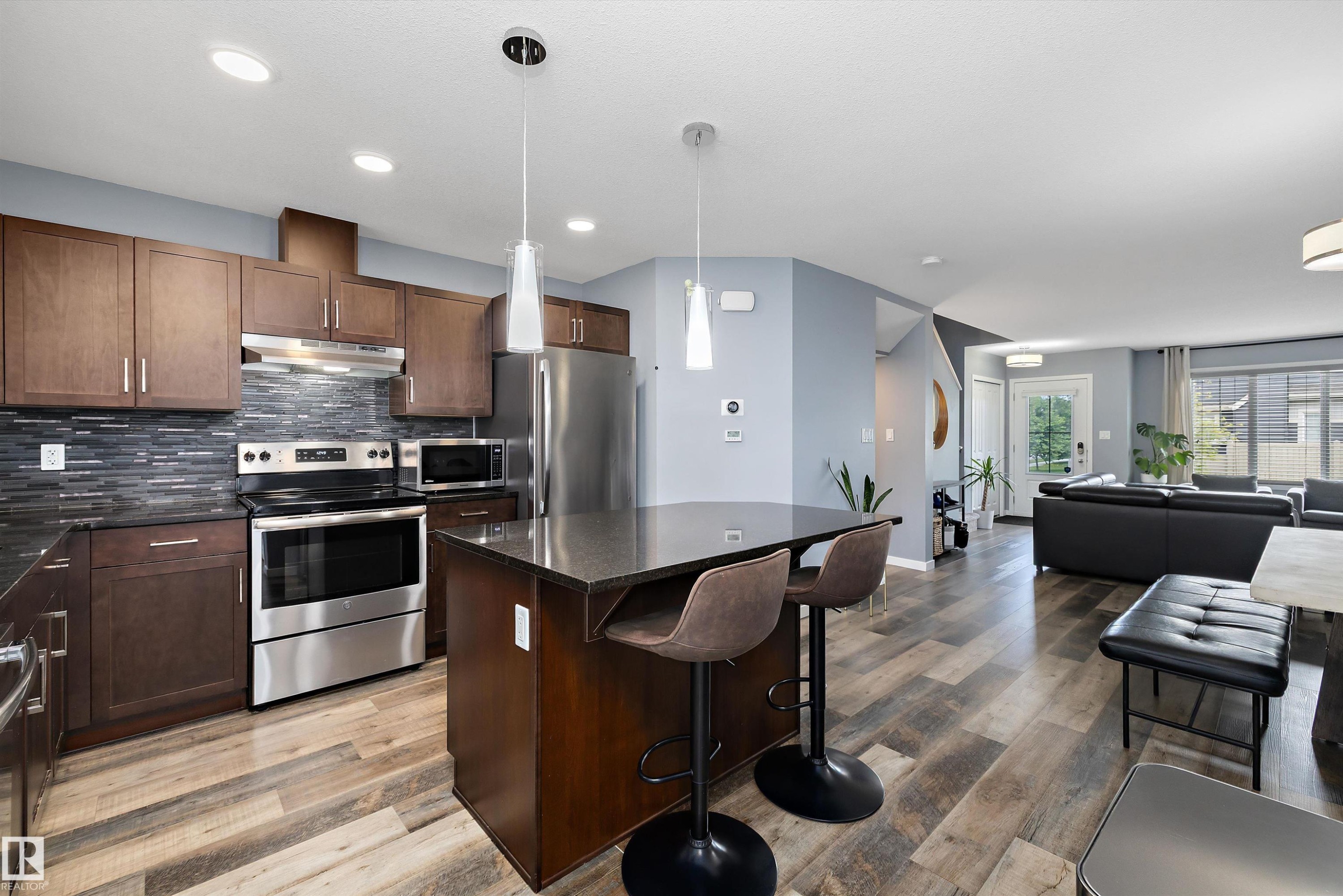 Open-concept kitchen featuring dark wood cabinetry, stainless steel appliances, a dark stone island with pendant lighting, a dark mosaic tile backsplash, and wood-finish flooring - 2833 Chokecherry Place, Edmonton, AB