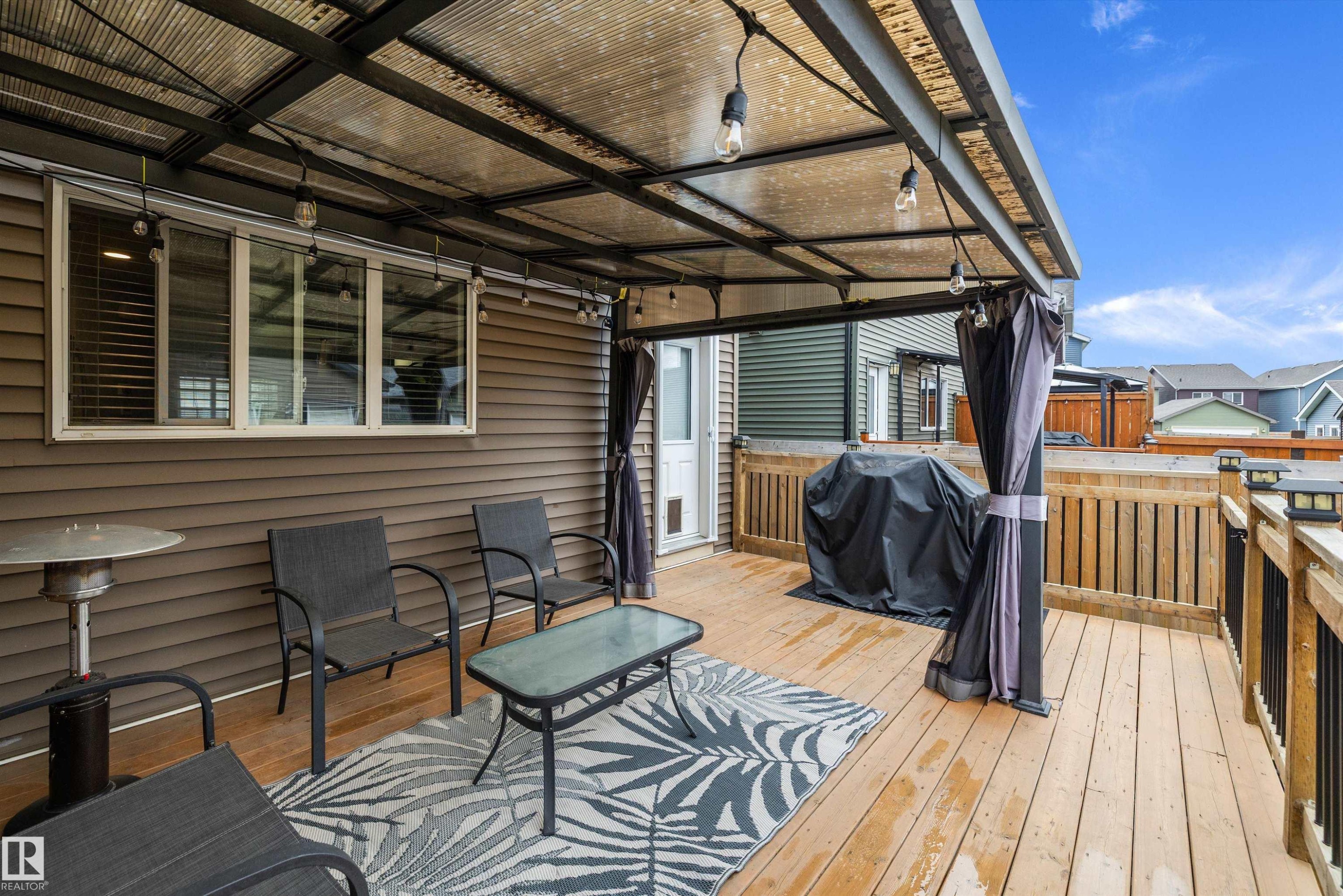 Covered outdoor deck with wood-finish flooring, featuring a translucent roof, string lighting, and wood railing with black metal spindles - 2833 Chokecherry Place, Edmonton, AB