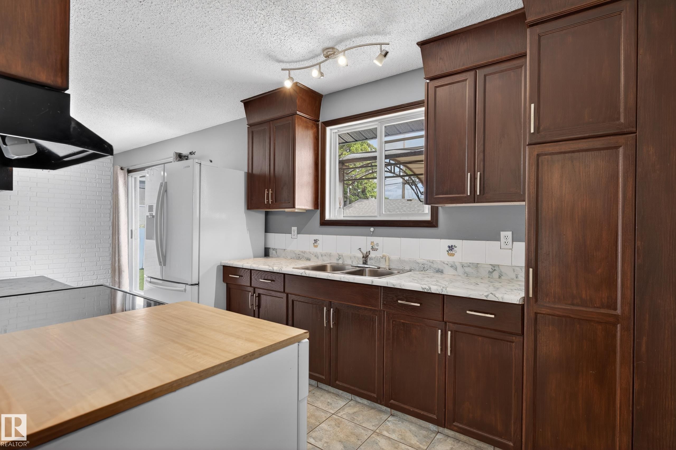 Kitchen featuring dark wood-finish cabinetry, light-toned countertops, a stainless steel double-basin sink, and a white tiled backsplash - 13427 114 Street Nw, Edmonton, AB