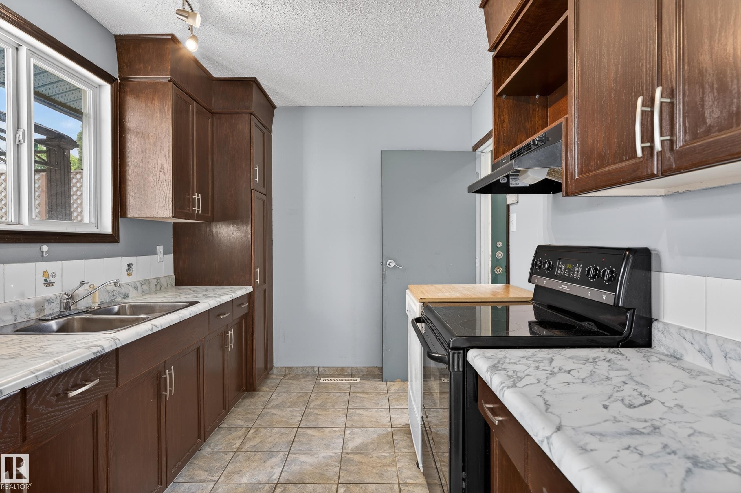 Kitchen featuring dark wood cabinetry, light-toned countertops with veining, a stainless steel double sink, a black electric range with an overhead hood, and tile flooring - 13427 114 Street Nw, Edmonton, AB
