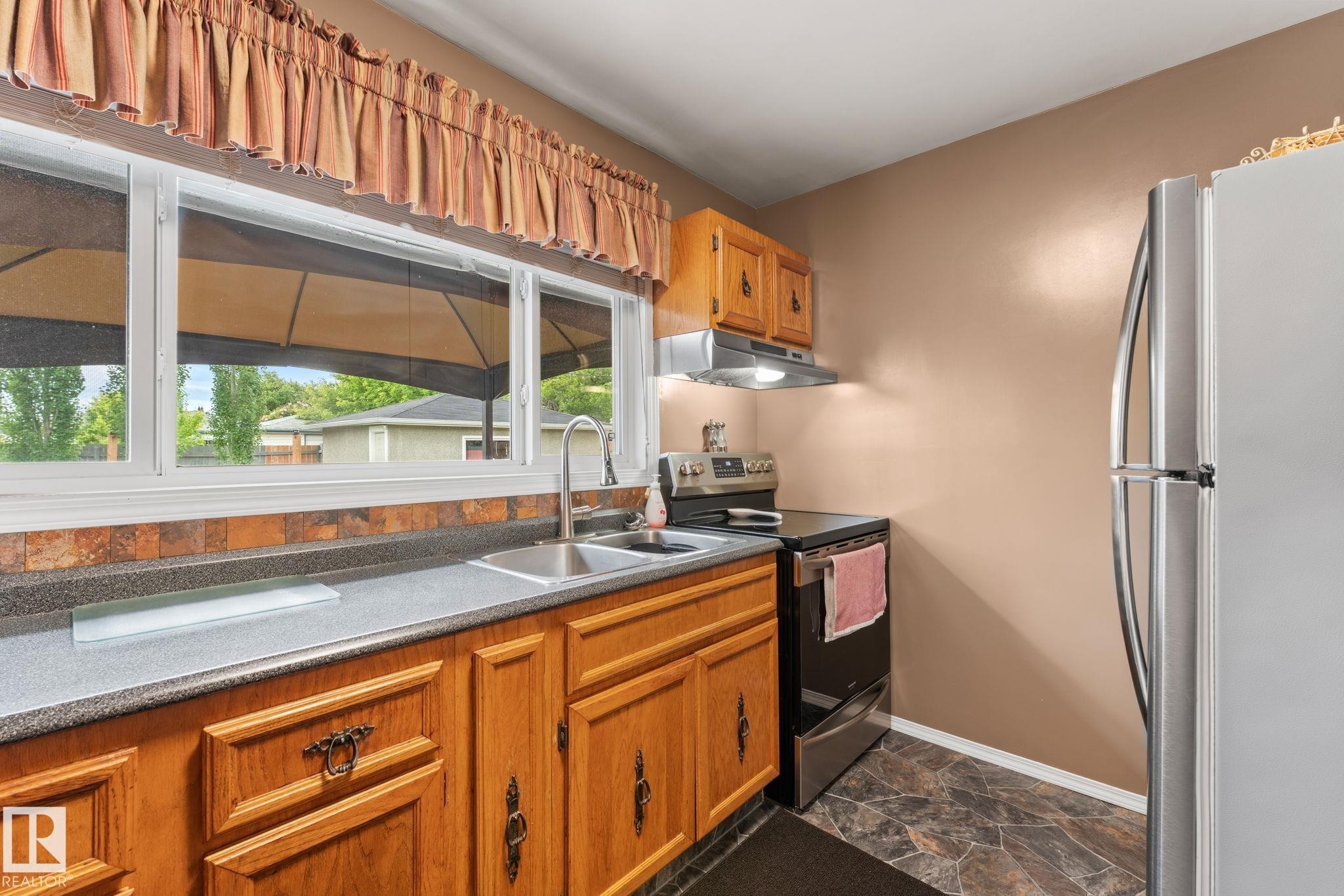 Kitchen featuring wood cabinetry, stone-look backsplash, and a stainless-steel sink with a gooseneck faucet - 11219 134A Avenue, Edmonton, AB