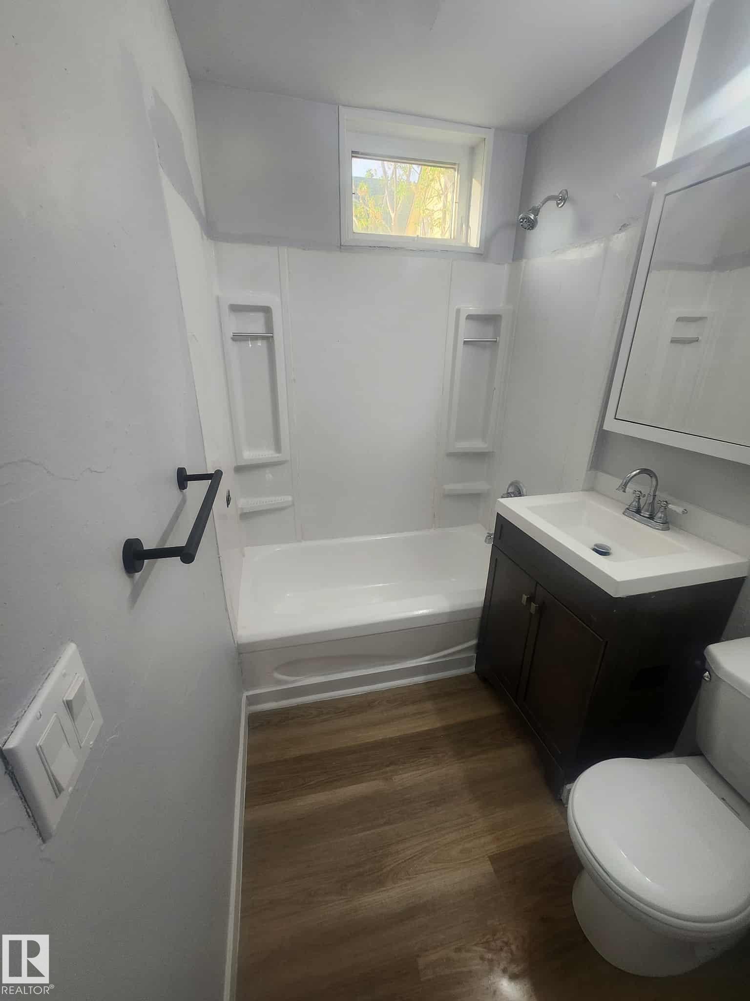 Bathroom featuring wood-finish flooring, a white bathtub with shower surround, a dark wood-finish vanity with an integrated sink, a wall-mounted mirror, and a window - 11308 94 Street, Edmonton, AB