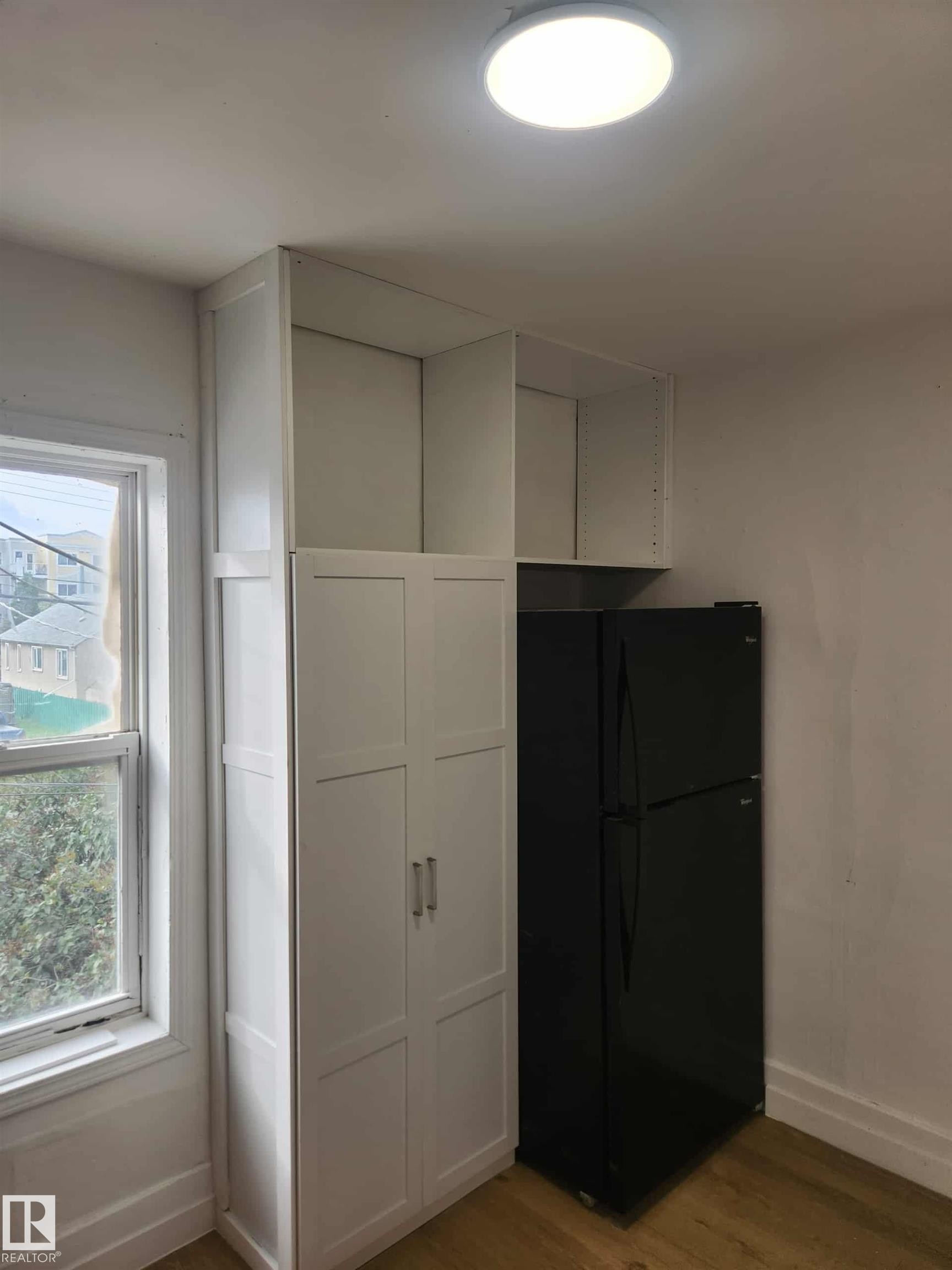 Kitchen area featuring a black refrigerator, tall white pantry cabinetry with silver hardware, upper open shelving, and wood-finish flooring - 11308 94 Street, Edmonton, AB