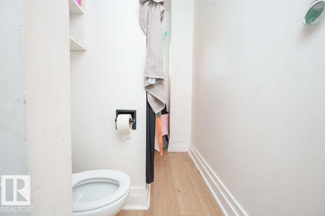 Compact water closet featuring a white toilet, light wood-finish flooring, and white wall shelving - 11308 94 Street, Edmonton, AB