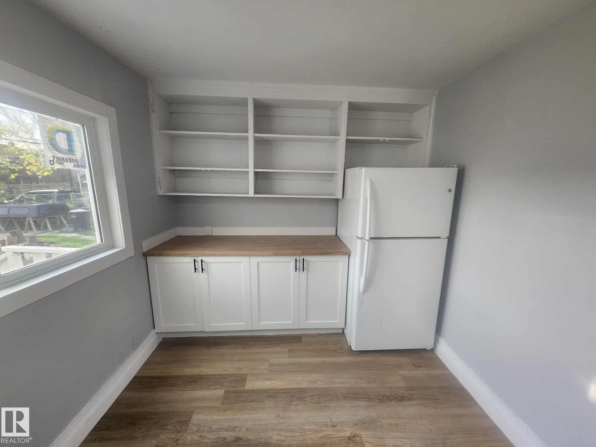 Compact kitchen area featuring white cabinetry with dark hardware, wood-finish countertops, open shelving, and a prominent window - 11308 94 Street, Edmonton, AB