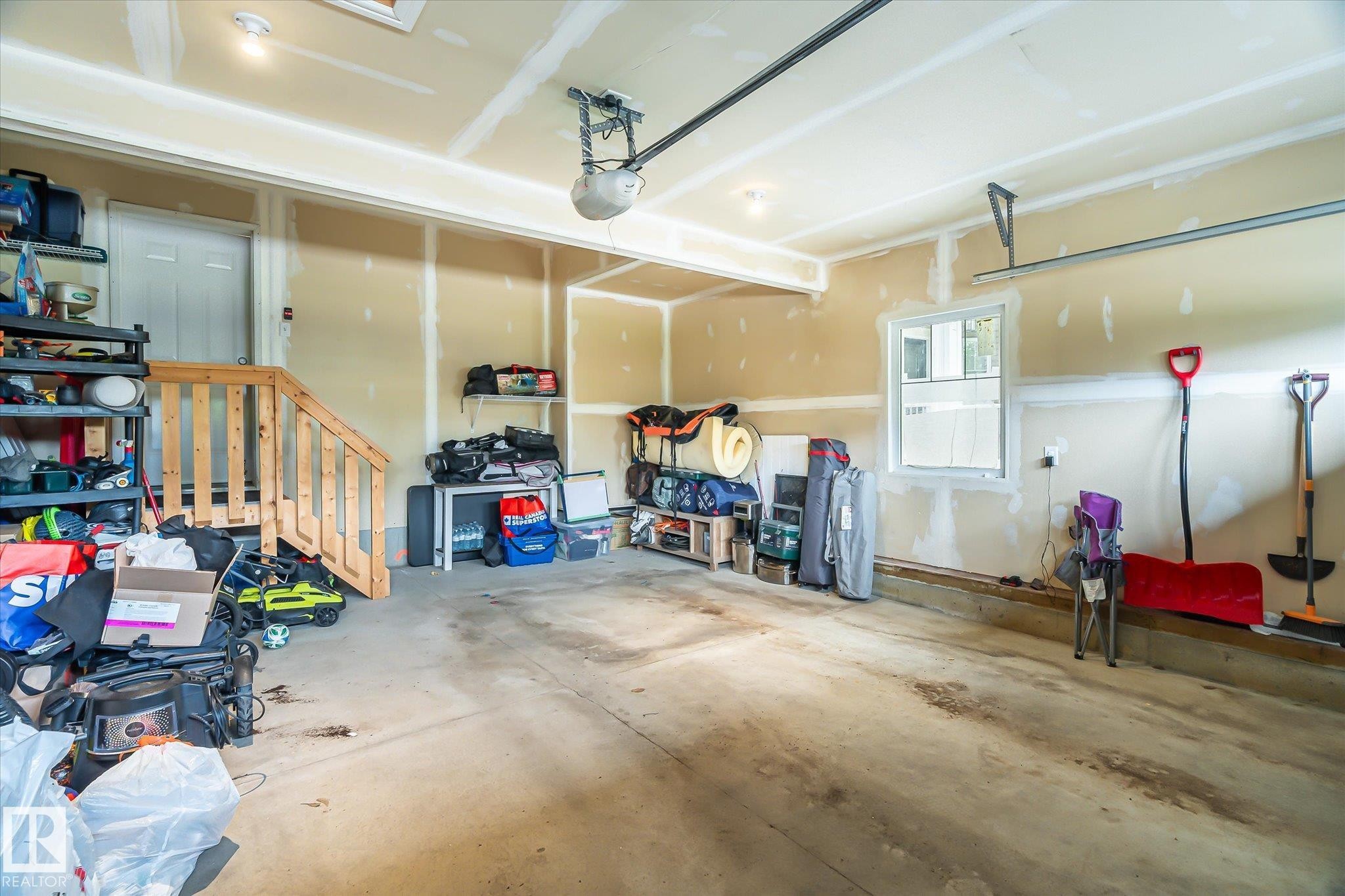 Attached garage featuring an automatic door opener, an interior side door, a window for natural light, exposed drywall, and a wooden staircase - 3685 Westcliff Way, Edmonton, AB