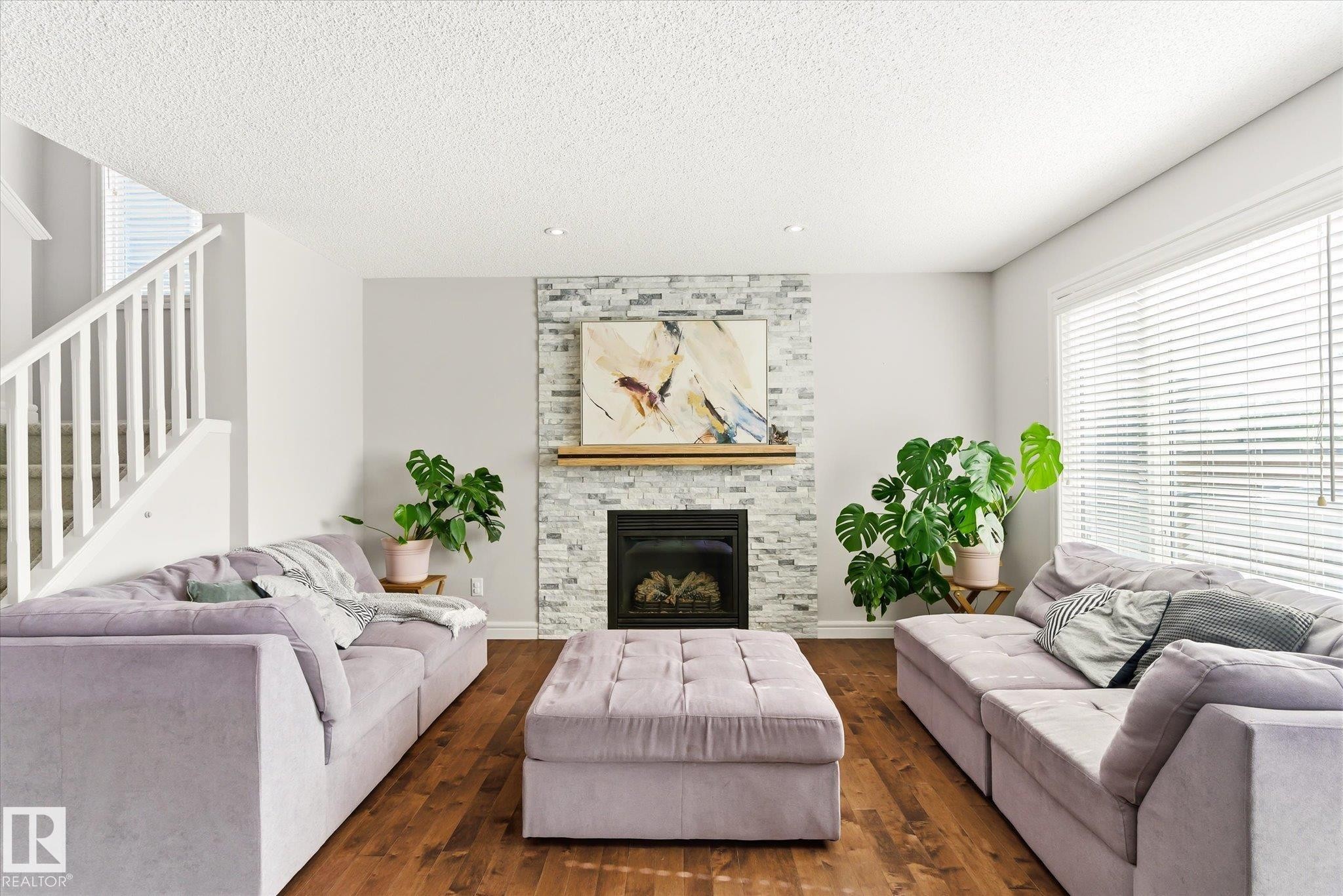 Spacious living area featuring a stacked stone fireplace with wood mantel, hardwood flooring, and a large window with horizontal blinds - 3685 Westcliff Way, Edmonton, AB