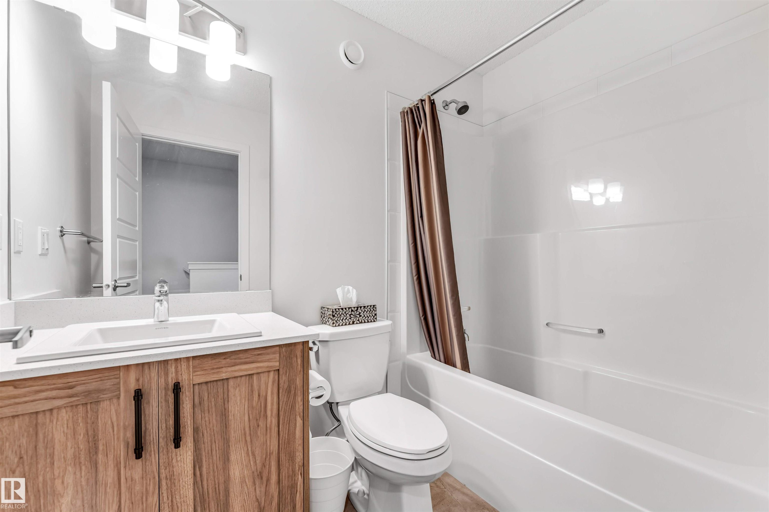 Bathroom featuring a wood-finish vanity with a white countertop and integrated sink, a large wall mirror, and a white tub/shower combination with a single shower head - 116 3305 Orchards Link Sw, Edmonton, AB