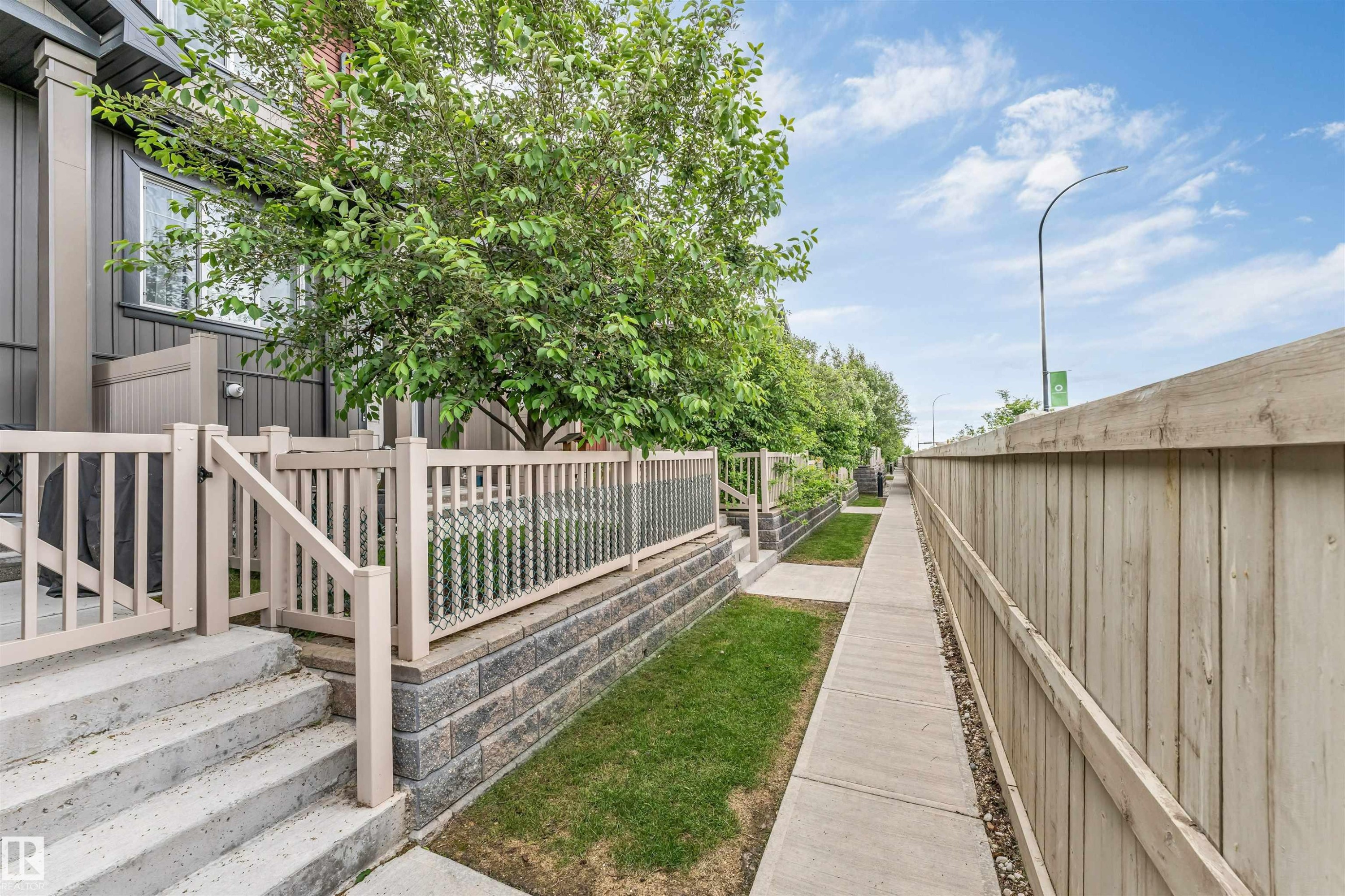 Exterior walkway featuring concrete paving, a tall wooden fence, and maintained grass areas - 116 3305 Orchards Link Sw, Edmonton, AB