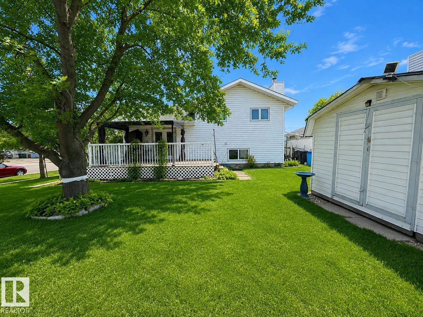Spacious backyard featuring a large wood-decked patio with white railing, a mature tree, a detached storage shed, and a stone garden border - 1 Munro Court, Leduc, AB