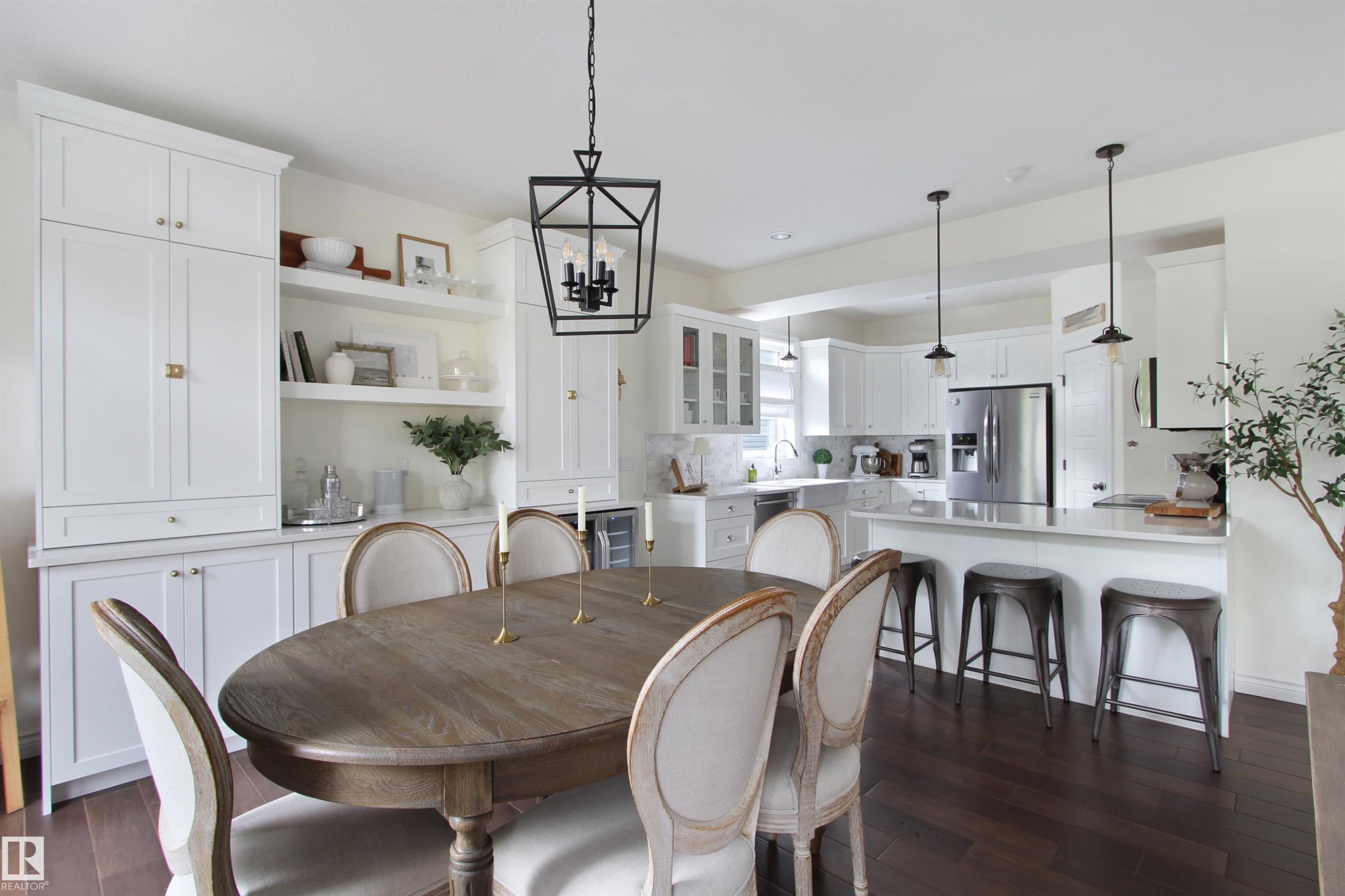 Open-concept kitchen and dining area featuring dark wood-finish flooring, a white kitchen island with seating, pendant lighting, stainless steel appliances, and white cabinetry - 34 Falcon Road, Cold Lake, AB
