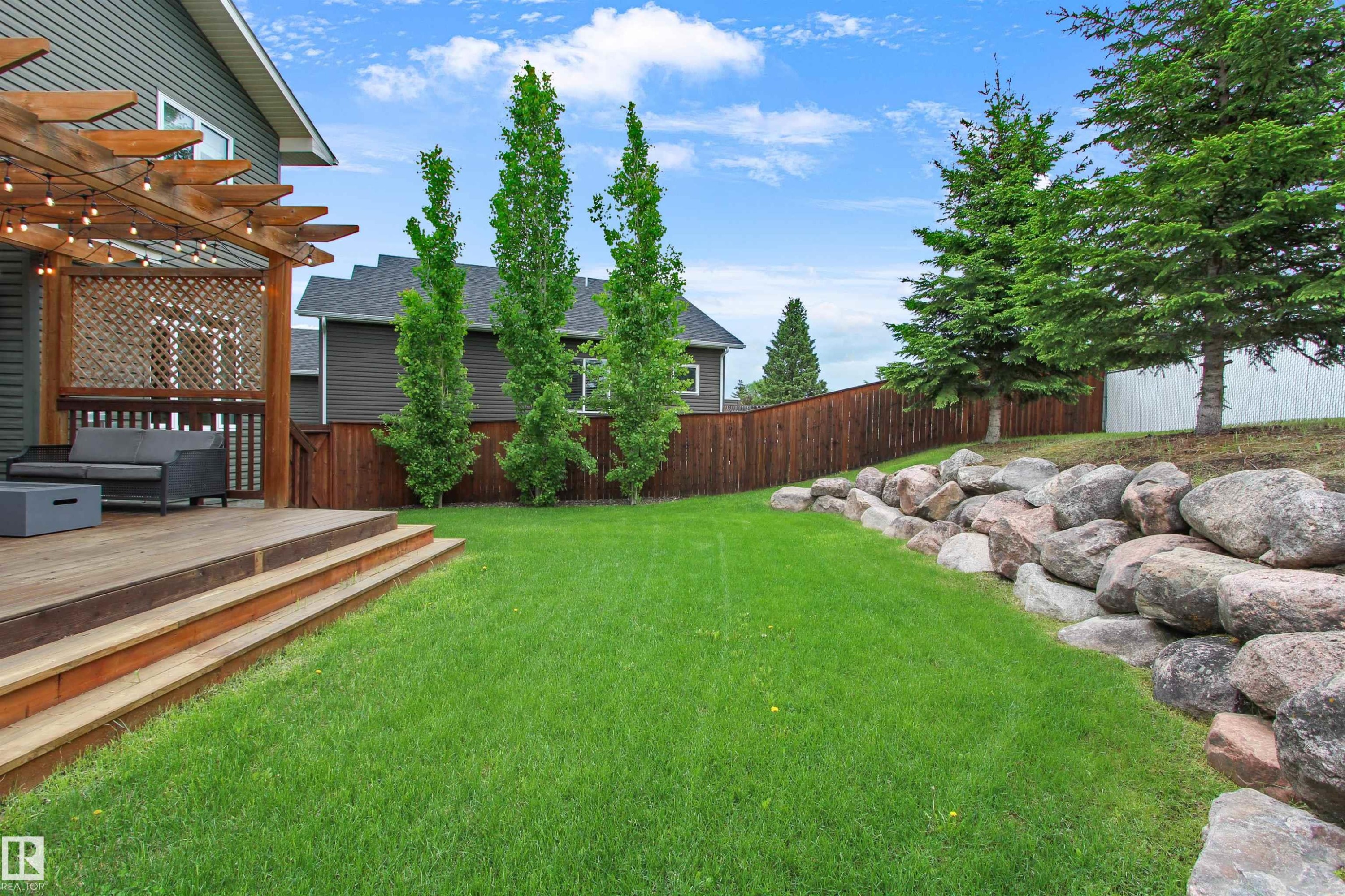 Expansive backyard featuring a large wood-finish deck with a pergola, tiered wood-finish steps, a rock retaining wall, a manicured lawn, and a privacy fence - 34 Falcon Road, Cold Lake, AB