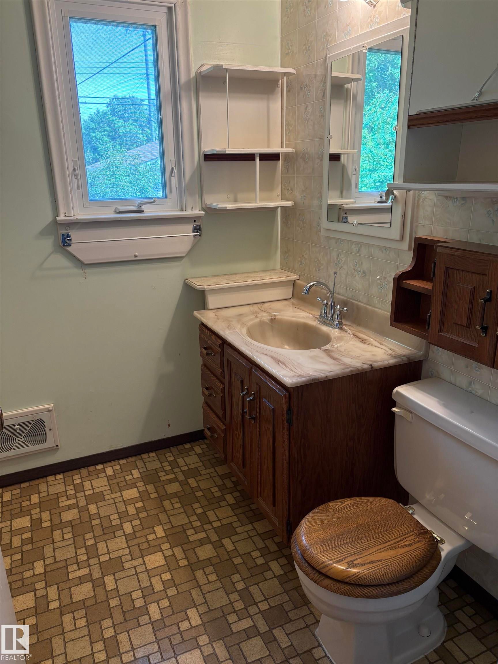 Bathroom featuring a window, built-in storage shelving, wood-finish vanity with an integrated sink, patterned floor tiling, and a toilet with a wood-finish seat - 5511 50 St, Tofield, AB