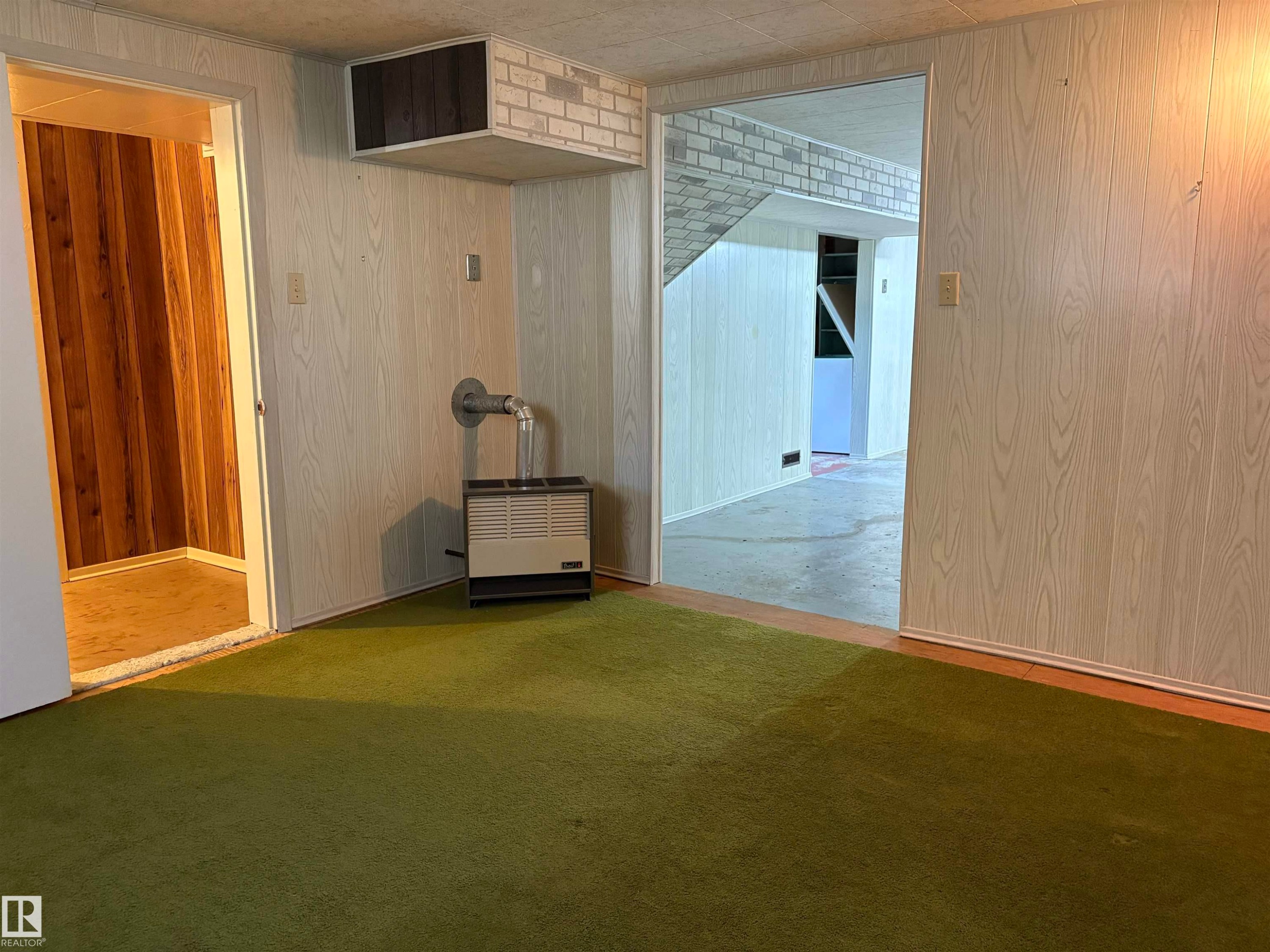 Interior space featuring green carpet flooring, light wood-grain paneling, a wall-mounted heating unit, and an overhead bulkhead with white brick detailing - 5511 50 St, Tofield, AB