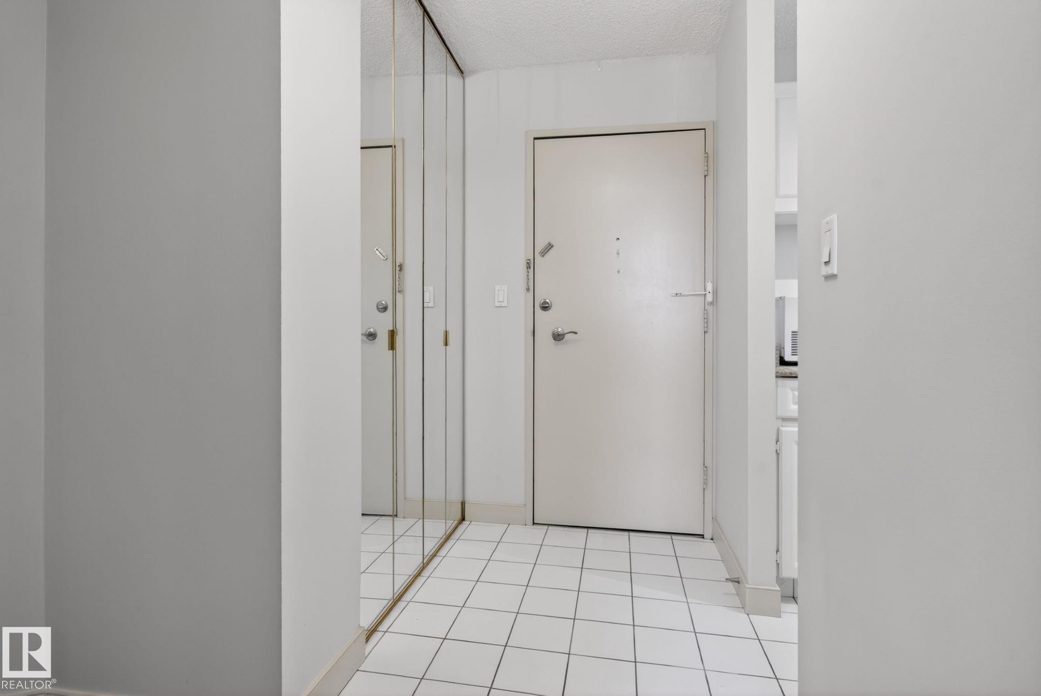 Entryway featuring square white floor tiles, a solid core entry door with polished chrome hardware, and a full-height mirrored wall - #412 13910 Stony Plain Rd Nw, Edmonton, AB - Indoor Photo Showing Other Room