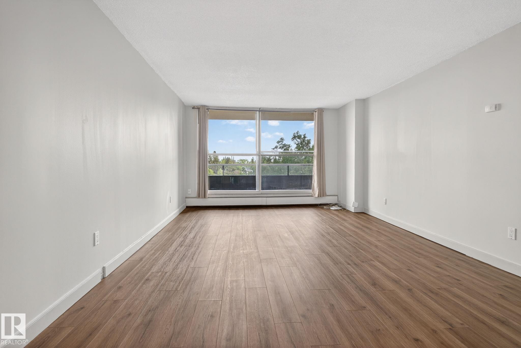 Expansive room featuring wood-finish flooring, white walls, and a large window with a garden outlook - #412 13910 Stony Plain Rd Nw, Edmonton, AB - Indoor Photo Showing Other Room