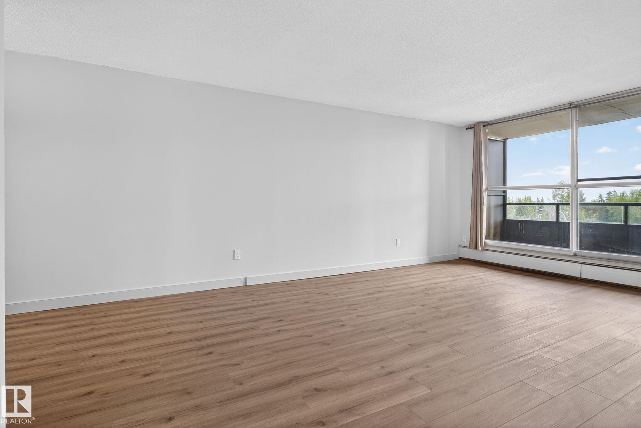 Spacious room featuring wood-finish flooring, white baseboards, and a large sliding glass door with a balcony access - #412 13910 Stony Plain Rd Nw, Edmonton, AB - Indoor Photo Showing Other Room