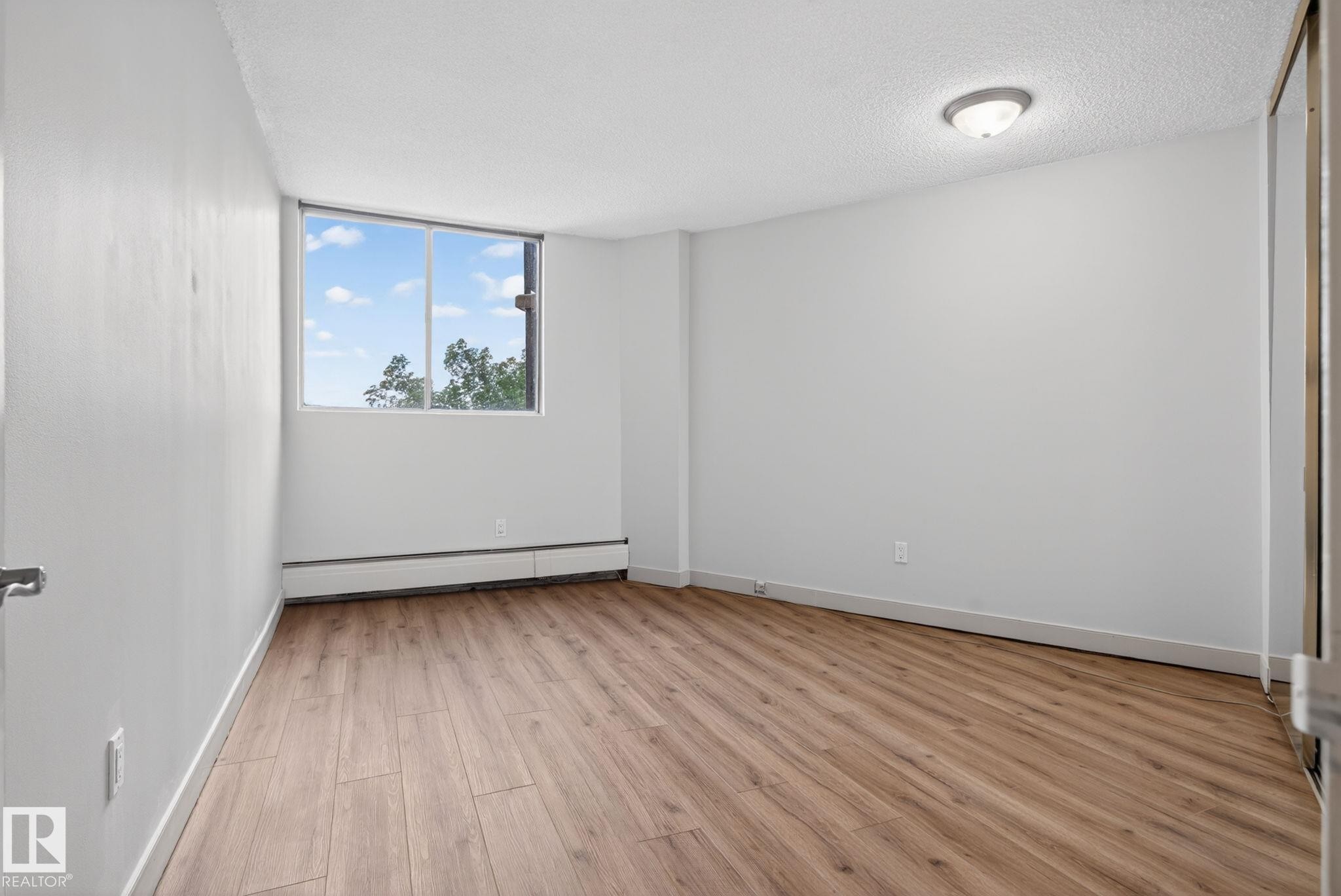 Bright room featuring a large window, wood-finish flooring, white painted walls, baseboard heating, and a flush-mount ceiling light - #412 13910 Stony Plain Rd Nw, Edmonton, AB - Indoor Photo Showing Other Room