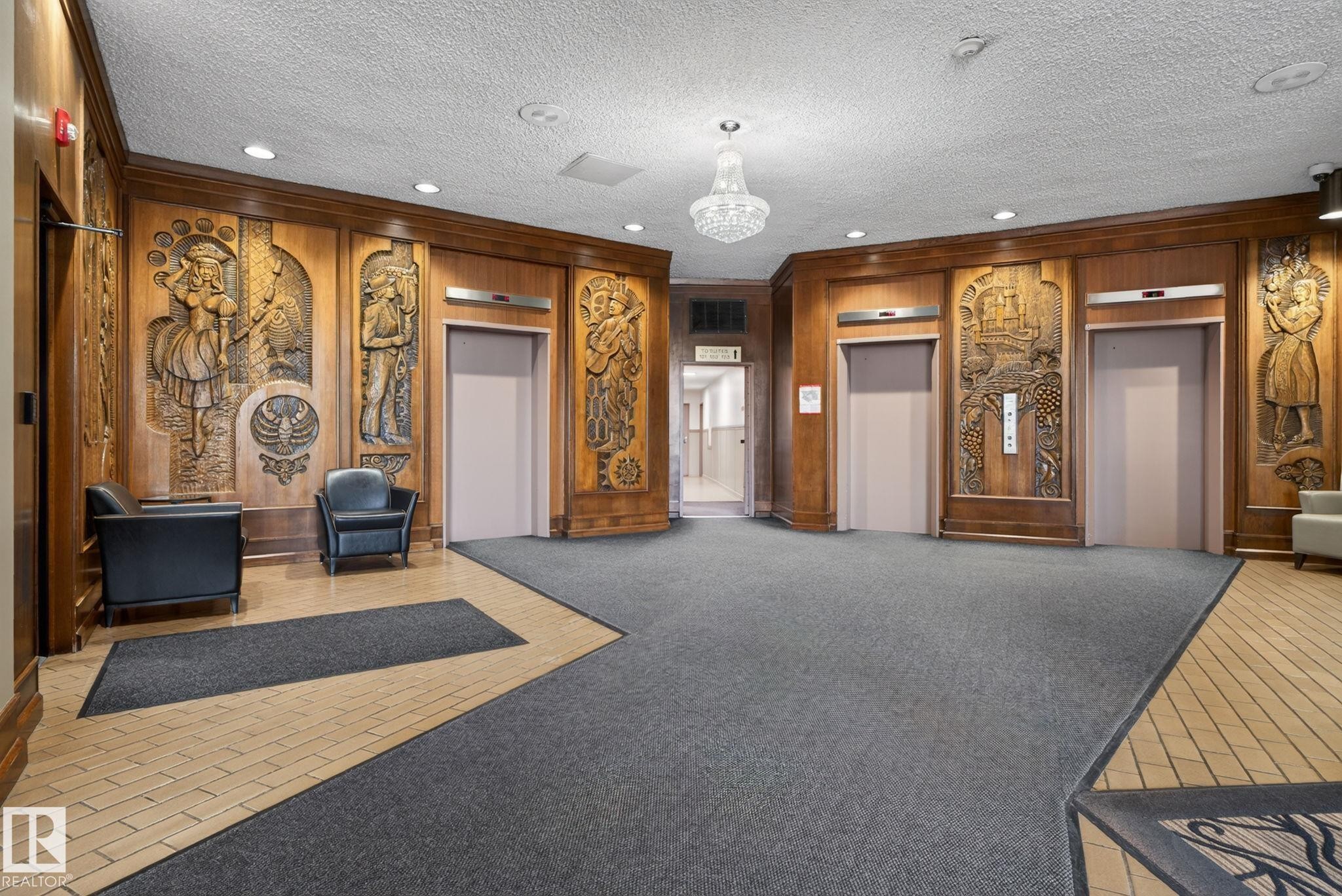 Elevator lobby featuring custom wood paneling with intricate carvings, a crystal chandelier, recessed lighting, and a combination of carpet and tile flooring - #412 13910 Stony Plain Rd Nw, Edmonton, AB - Indoor Photo Showing Other Room