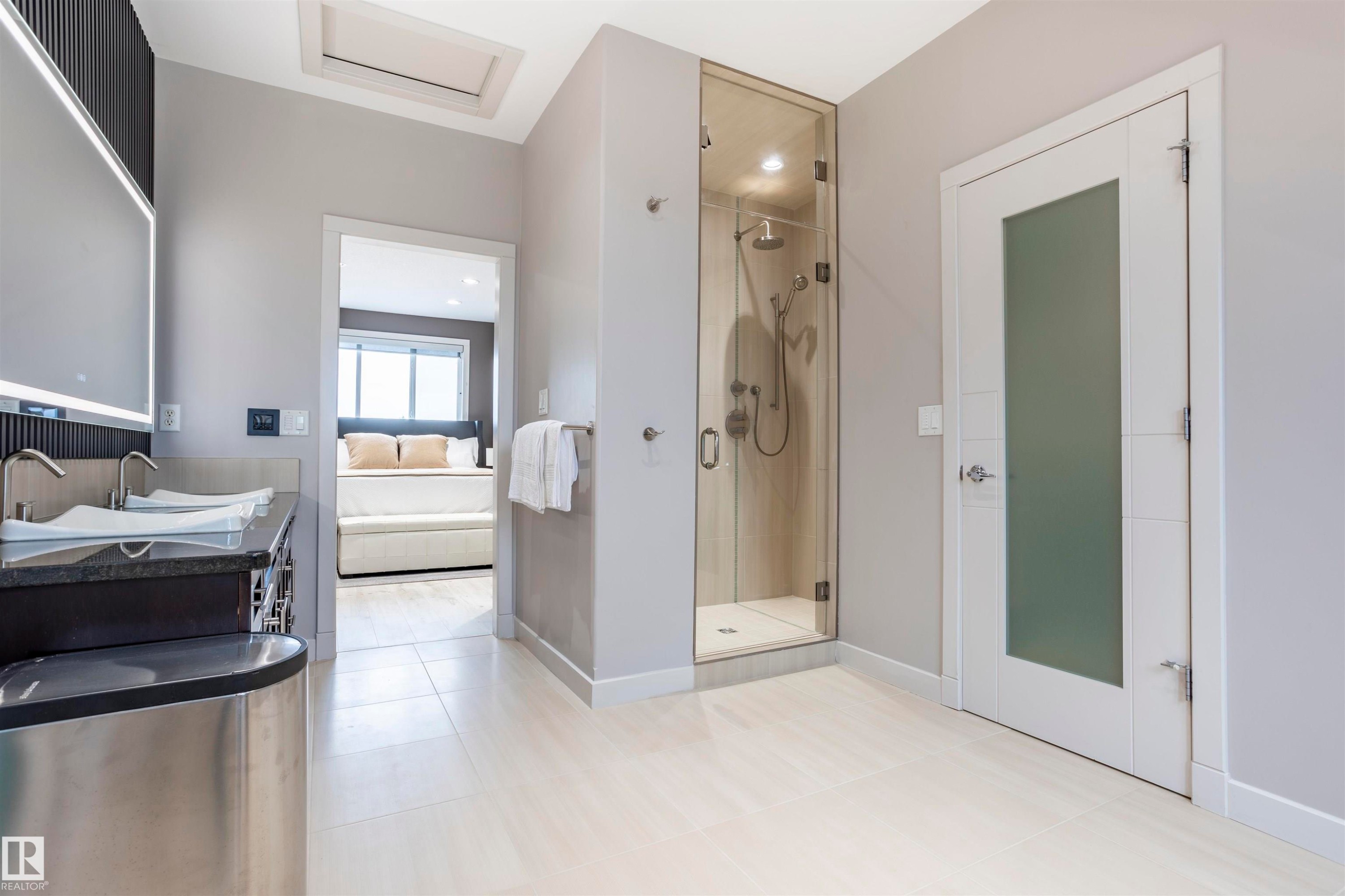 Contemporary bathroom featuring a glass-enclosed shower, dual vessel sinks on a dark countertop, an illuminated vanity mirror, and light tile flooring - 4949 Terwillegar Common Nw, Edmonton, AB