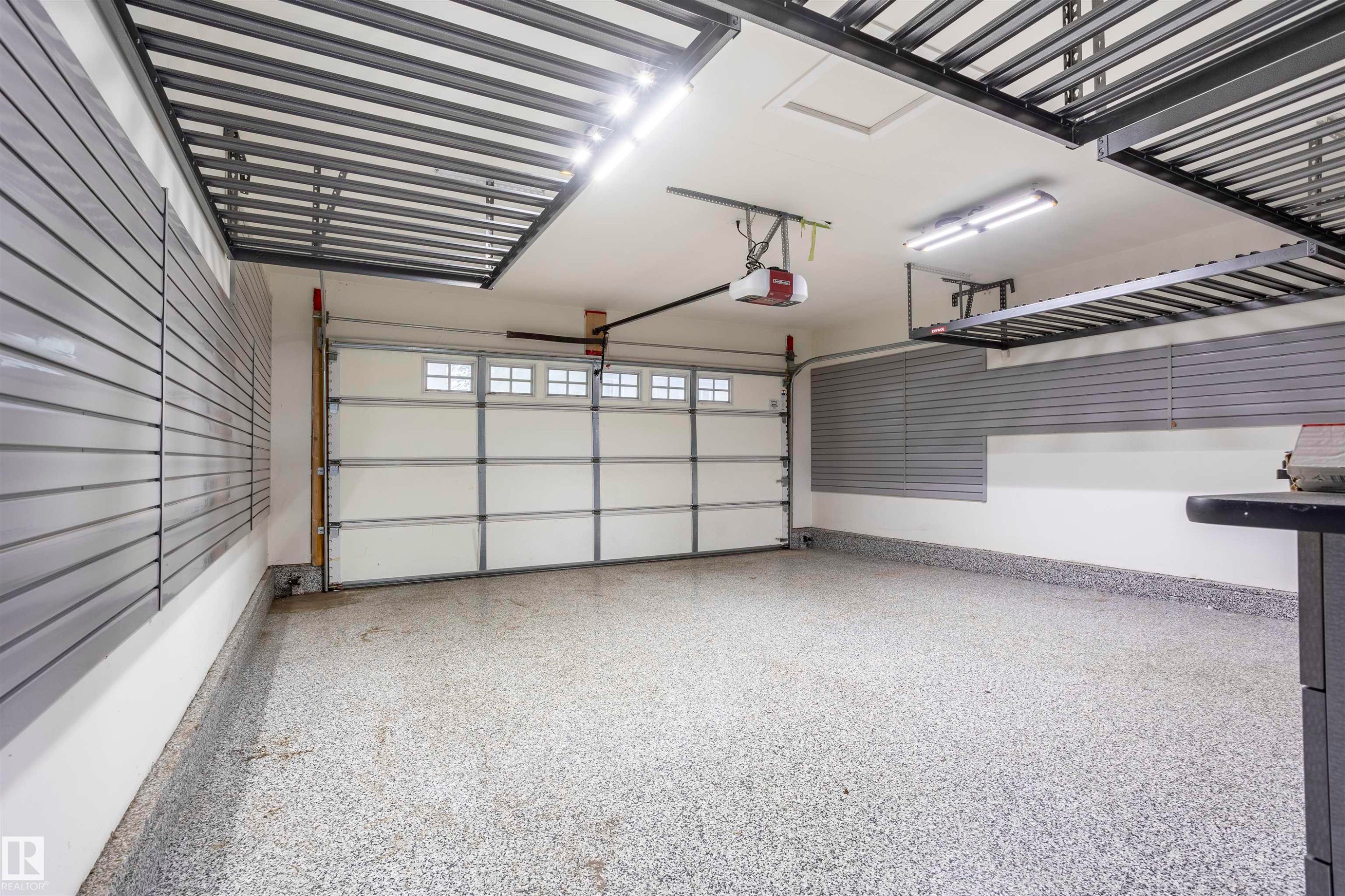 Garage featuring a speckled epoxy floor, overhead storage racks, slat wall organizational panels, and an automatic garage door with upper window inserts - 4949 Terwillegar Common Nw, Edmonton, AB