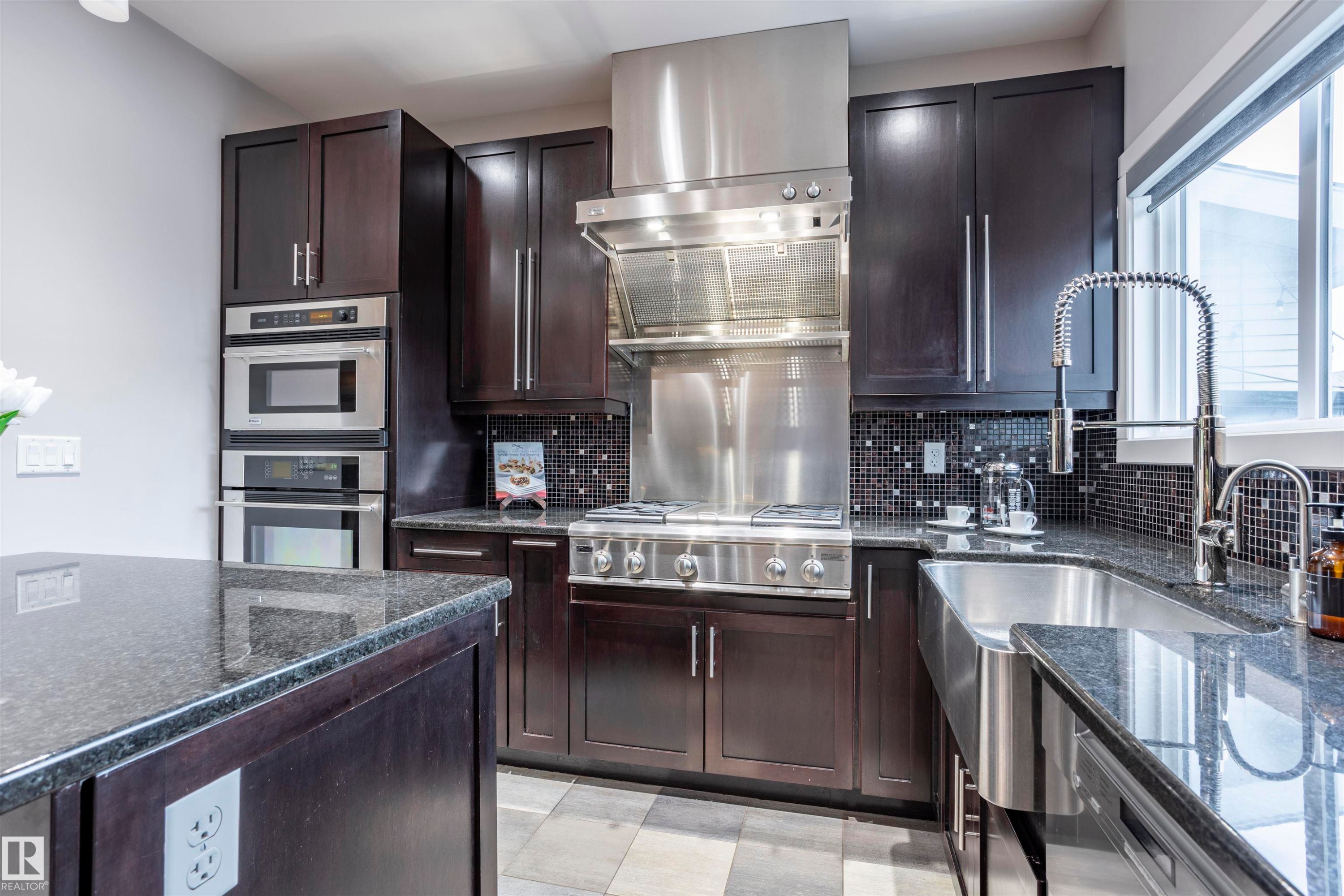 Gourmet kitchen featuring dark wood cabinetry, granite countertops, a built-in stainless steel microwave and oven, a stainless steel gas range with a matching range hood, and a farmhouse-style sink - 4949 Terwillegar Common Nw, Edmonton, AB