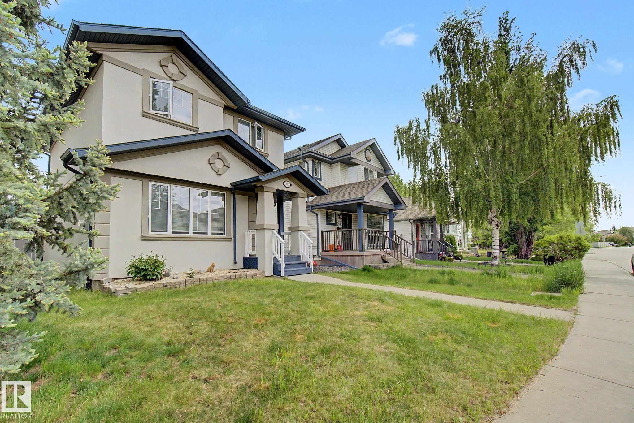 Stucco exterior with contrasting trim, gable rooflines, covered front porch with white railings, and mature landscaping - 710 Glenwright Court, Edmonton, AB