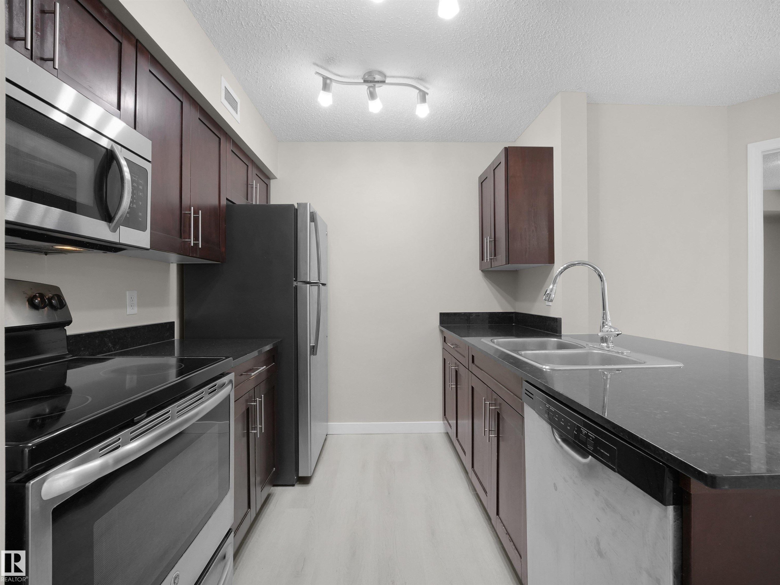 Galley kitchen featuring dark wood cabinetry, stainless steel appliances, black countertops, and a double basin sink - 220 12035 22 Avenue, Edmonton, AB