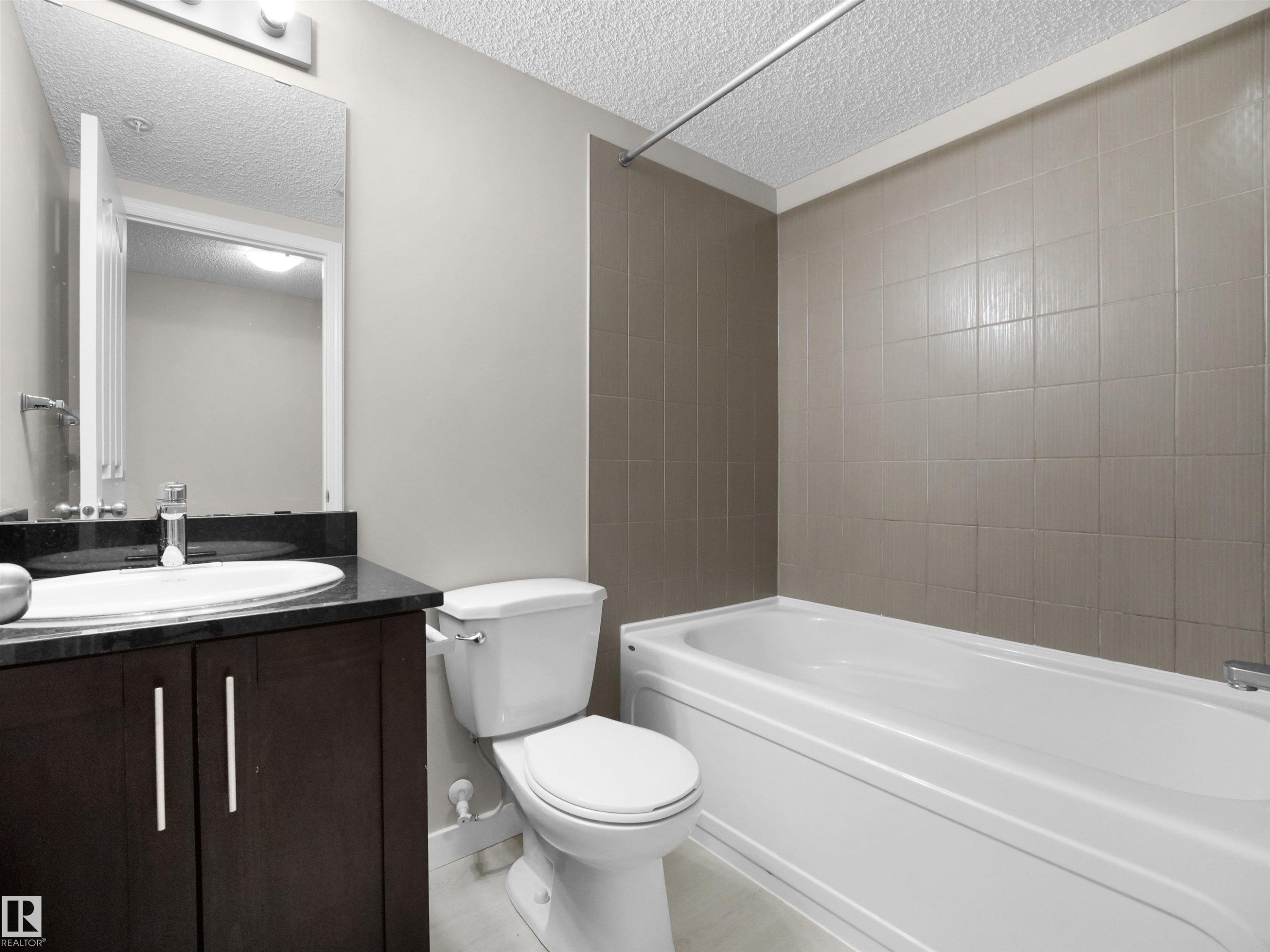 Bathroom featuring a white bathtub, tiled shower surround, dark wood-finish vanity with a black countertop, undermount sink, and a wall-mounted mirror - 220 12035 22 Avenue, Edmonton, AB