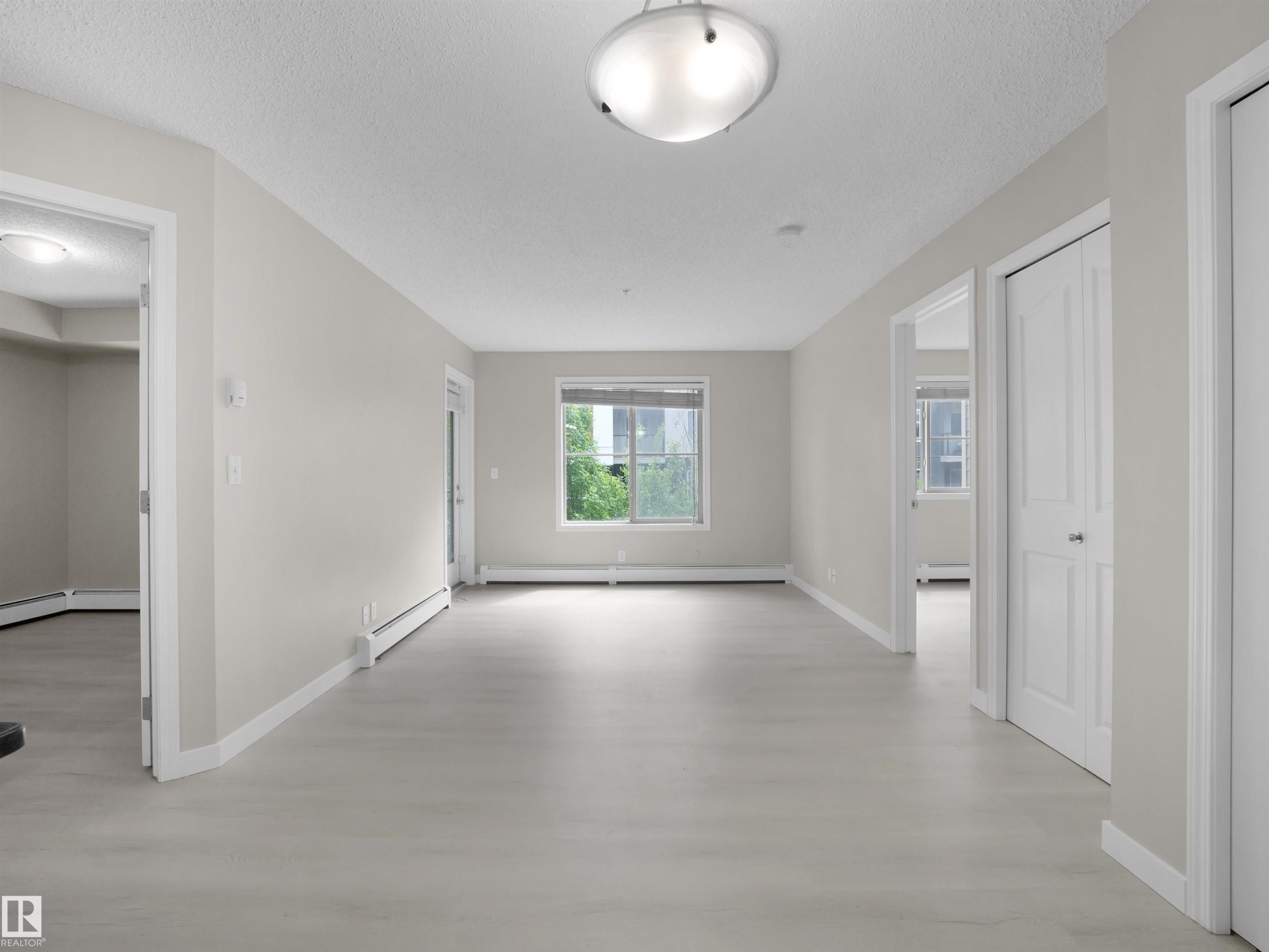 Spacious room featuring light-toned walls, wood-finish flooring, a window offering natural light, and a modern flush-mount ceiling light fixture - 220 12035 22 Avenue, Edmonton, AB