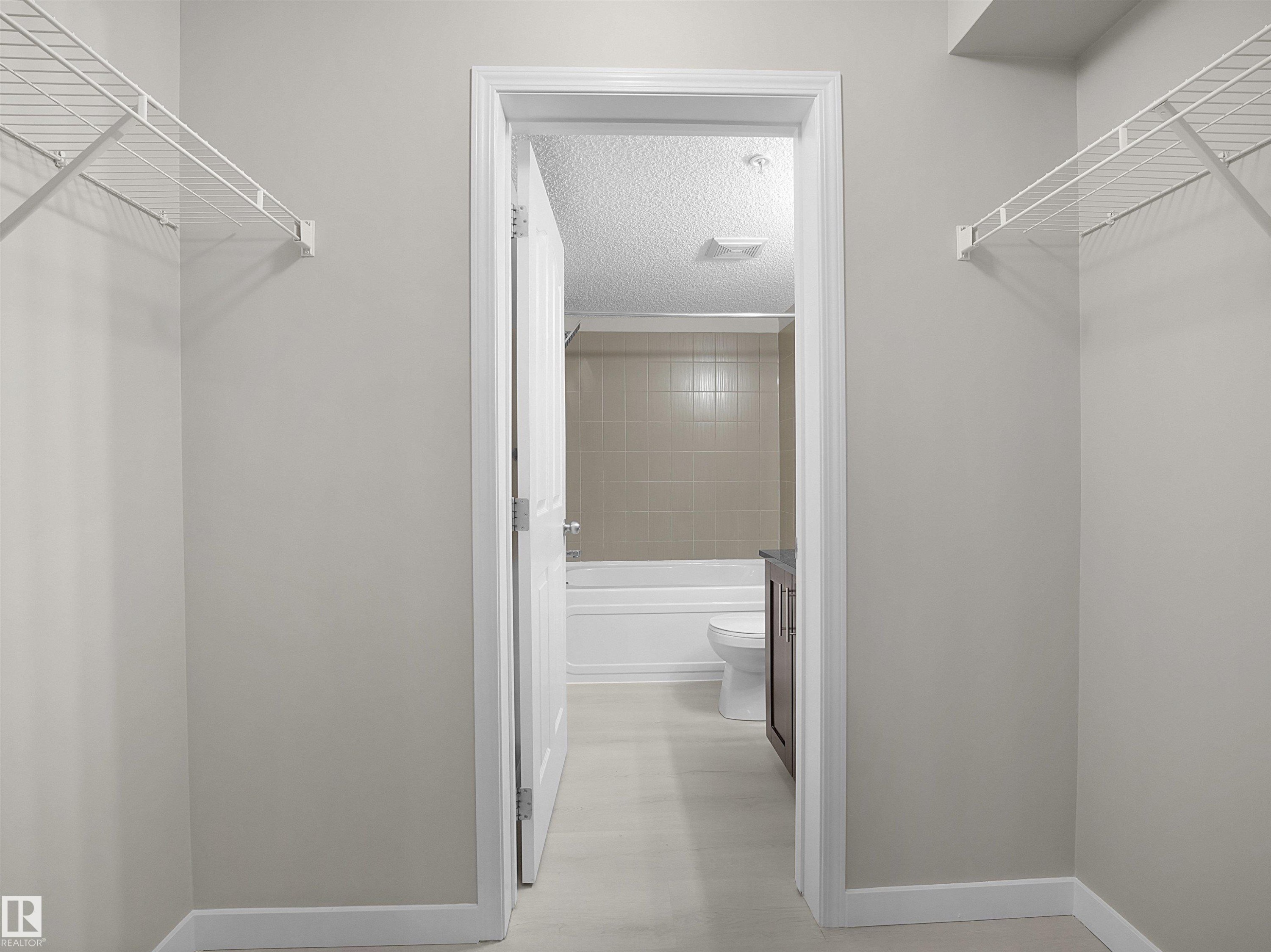 Walk-through closet with built-in wire shelving, neutral wall paint, and white trim, opening to a full bathroom featuring a white bathtub, tiled shower surround, and a dark wood-finish vanity - 220 12035 22 Avenue, Edmonton, AB