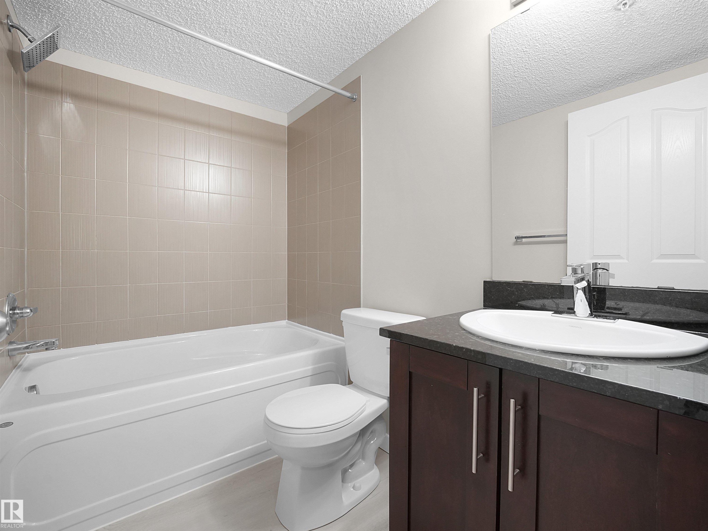 Bathroom featuring a white bathtub with an integrated shower, beige tiled surround, and a dark wood-finish vanity with a dark countertop and undermount sink - 220 12035 22 Avenue, Edmonton, AB