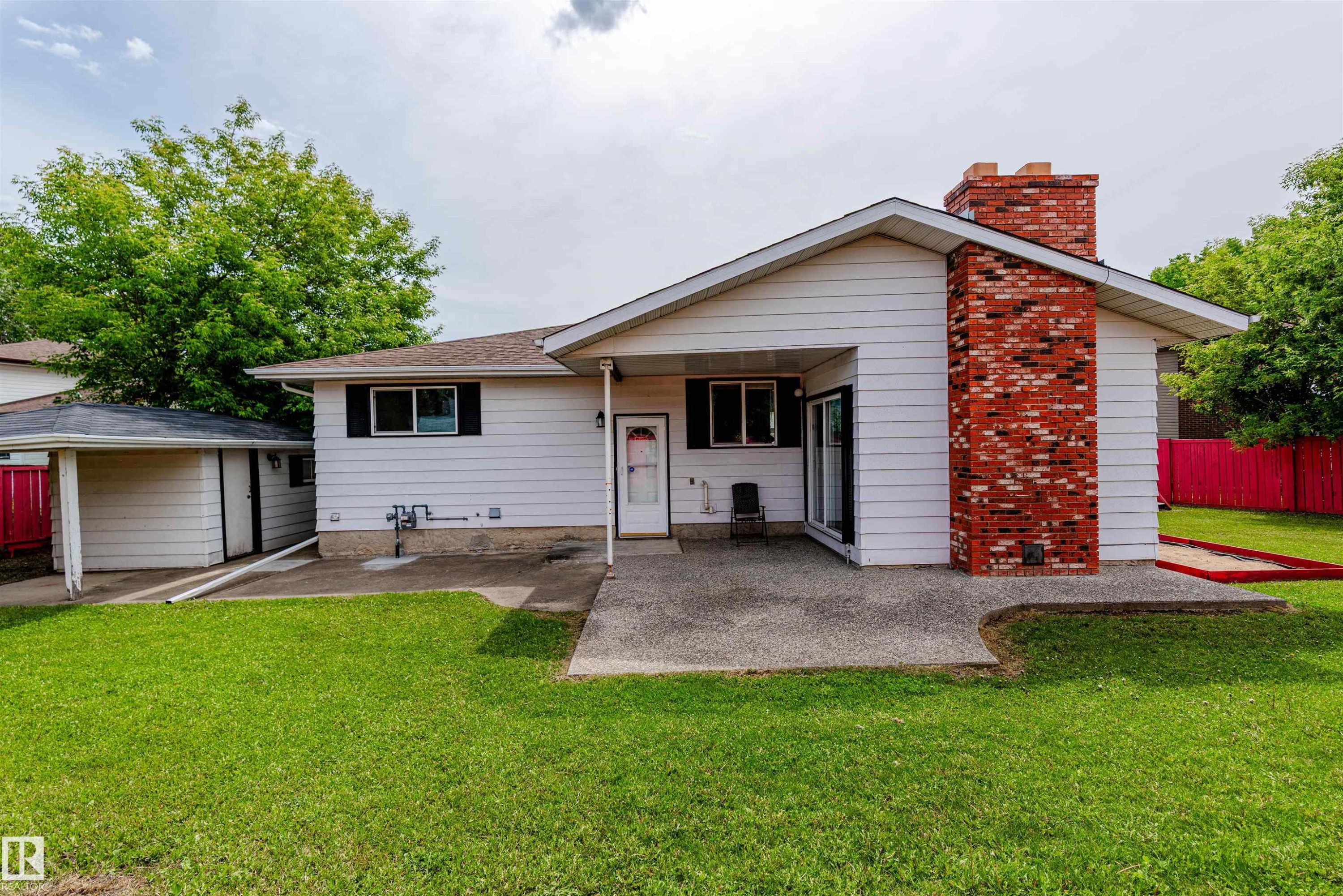 Rear exterior featuring white horizontal siding, a red brick chimney, a concrete patio, and a grass lawn - 16524 116 Street, Edmonton, AB