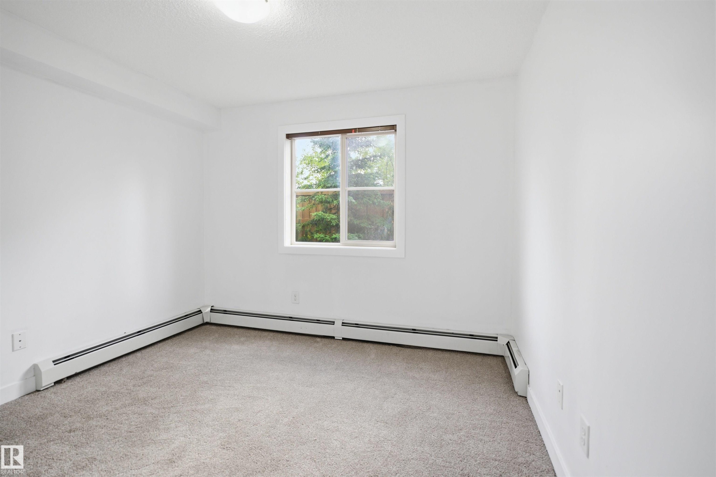 Bright room featuring a window with exterior tree scenery, light-colored carpeting, white baseboard heating units, and a ceiling-mounted light fixture - 115 274 Mcconachie Drive, Edmonton, AB