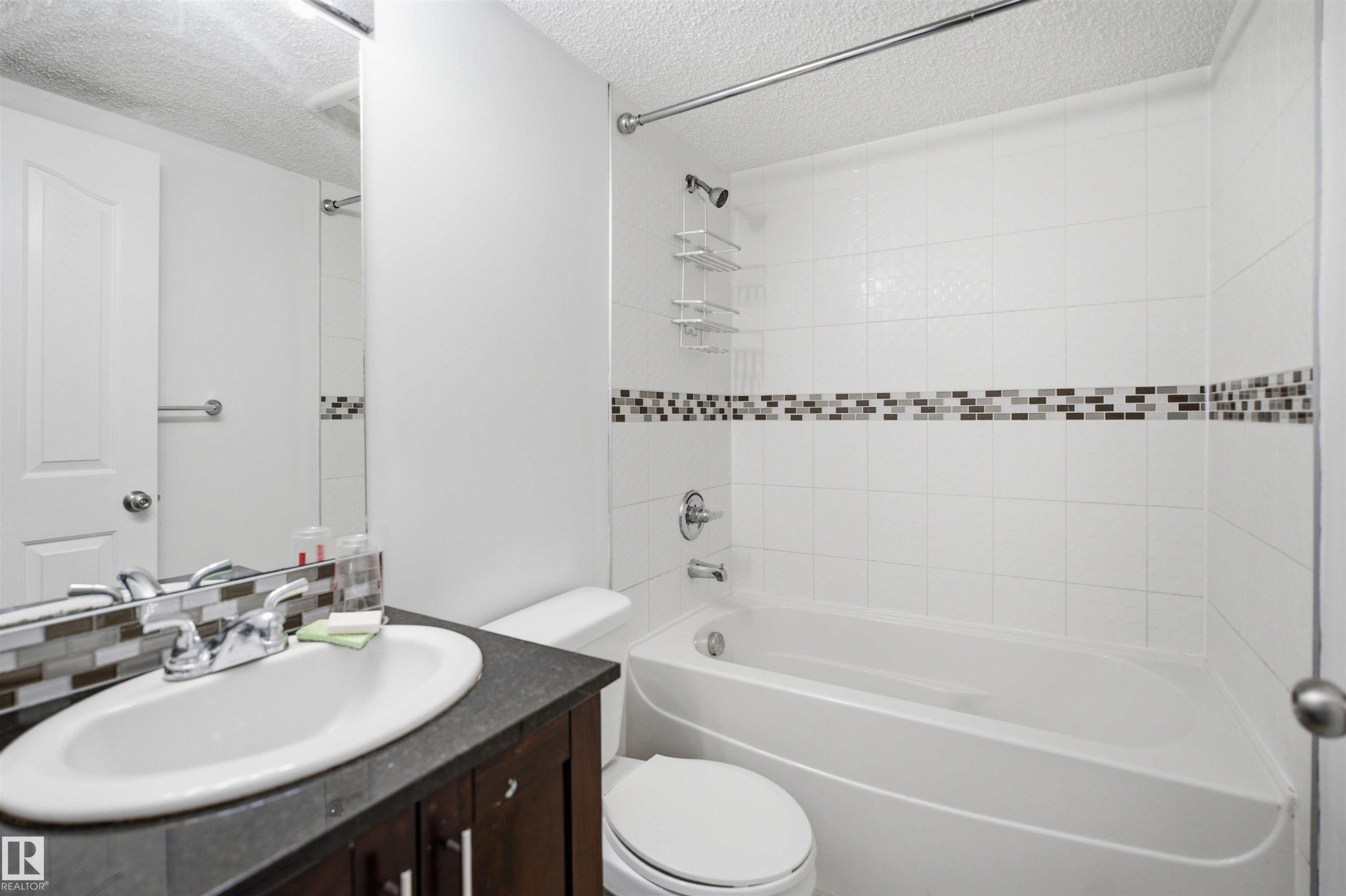 Bathroom featuring a white bathtub, white tiled shower surround with mosaic accent band, dark wood-finish vanity with an oval sink, and a large wall-mounted mirror - 115 274 Mcconachie Drive, Edmonton, AB