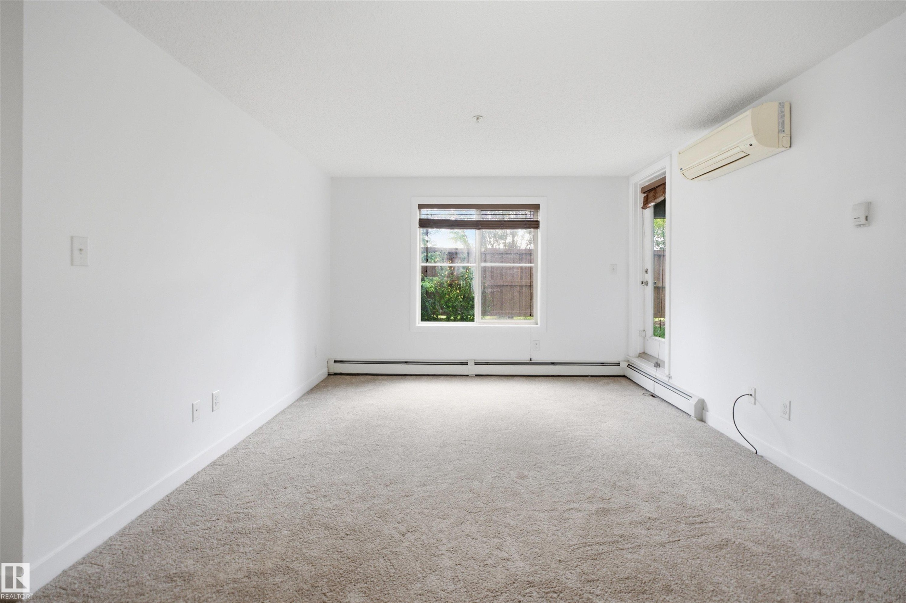 Spacious room featuring neutral carpeting, white walls, a window with blinds, a white door, and a wall-mounted air conditioning unit - 115 274 Mcconachie Drive, Edmonton, AB