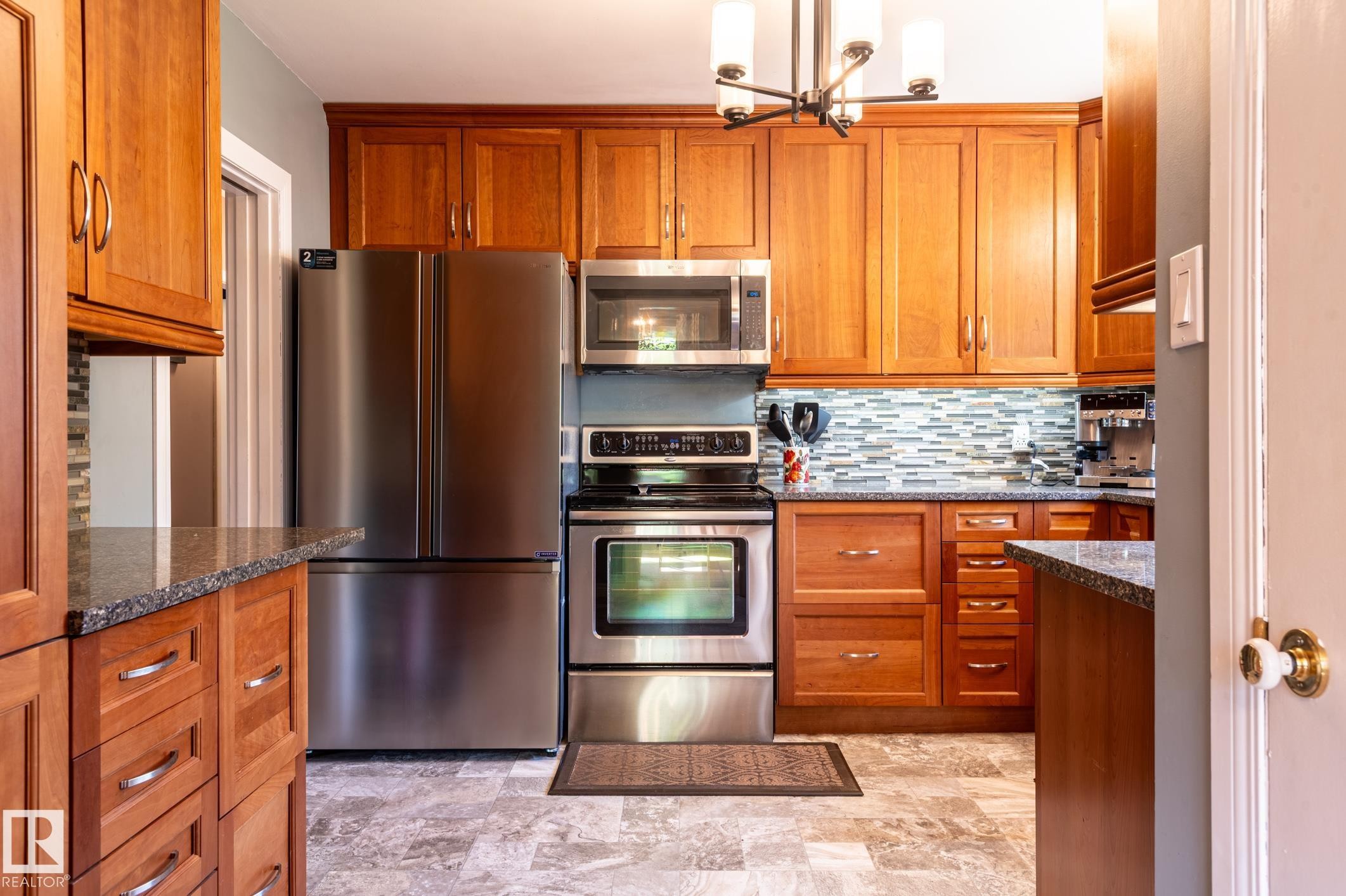 Kitchen featuring wood cabinetry, stainless steel appliances, speckled stone countertops, a mosaic tile backsplash, and light-toned floor tile - 14708 108 Av, Edmonton, AB