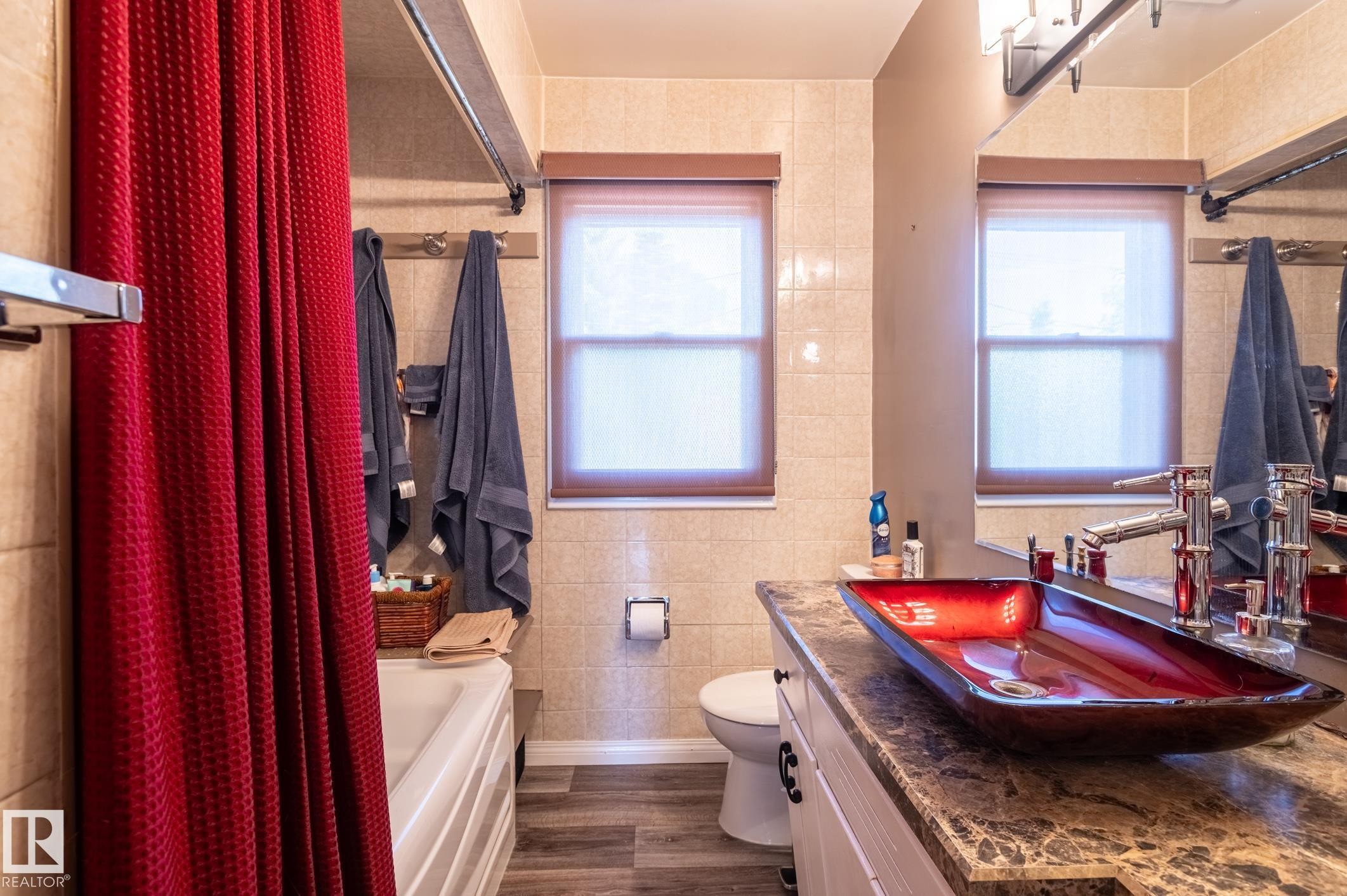 Bathroom featuring a vessel sink with a dark red finish, a vanity with a stone-finish countertop, wood-finish flooring, tiled walls, and two windows with roller blinds - 14708 108 Av, Edmonton, AB