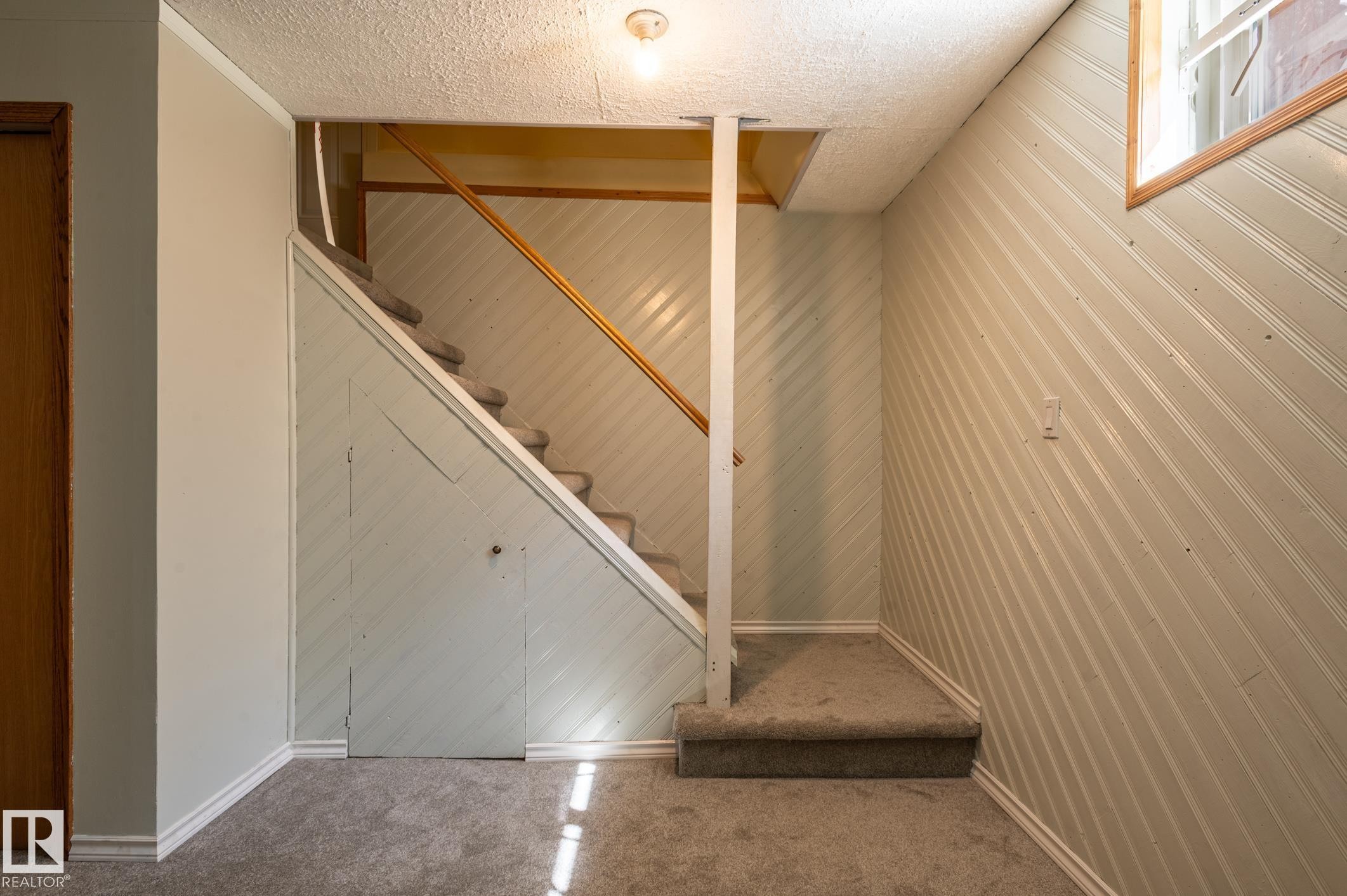 Carpeted staircase with a wood handrail, featuring light-colored diagonal paneling, a wooden interior door, and a built-in storage area under the stairs - 14708 108 Av, Edmonton, AB