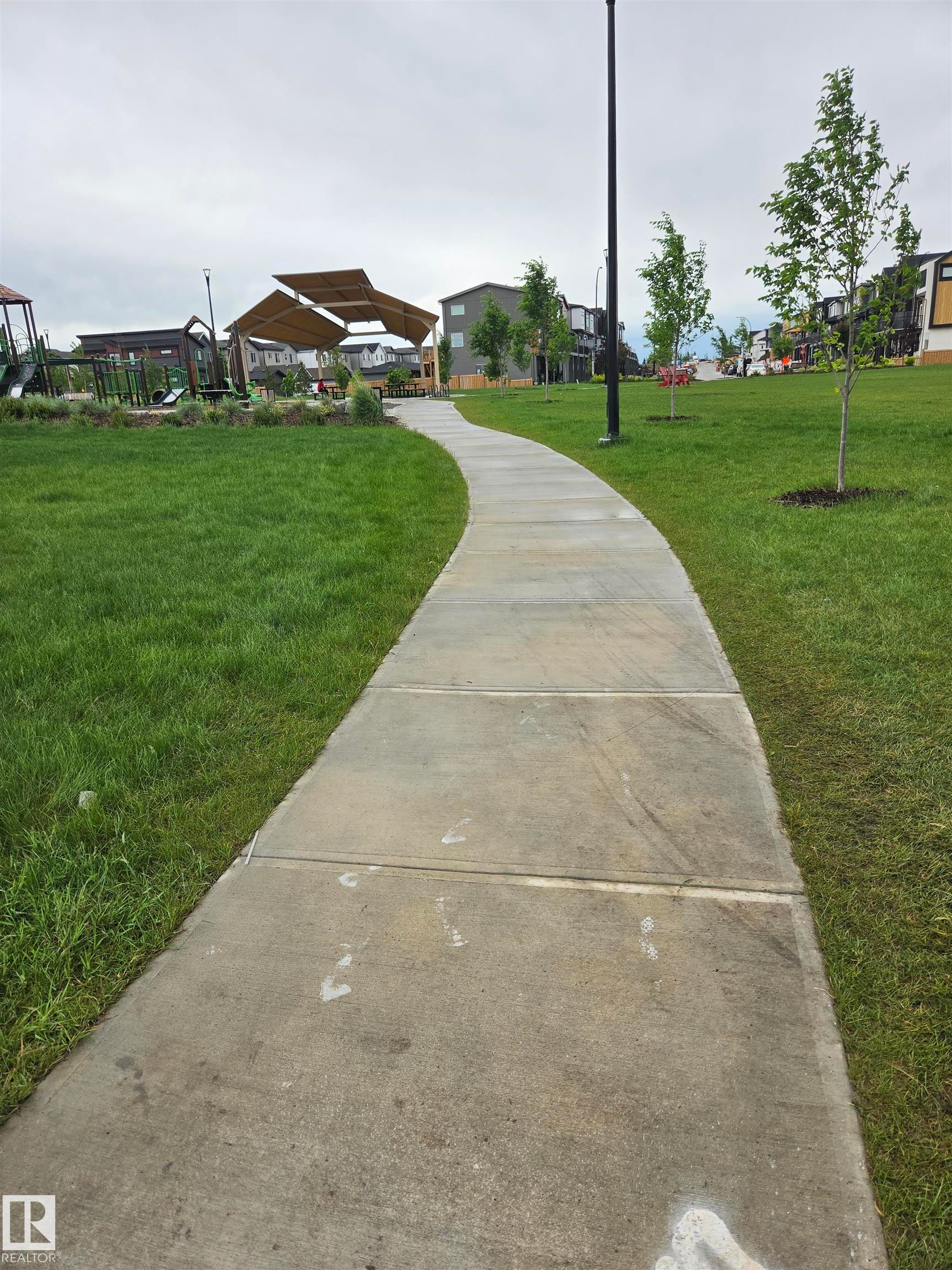 Curved concrete pathway bordered by manicured green lawns, leading towards a shaded seating area and play structure - 113 29A Street, Edmonton, AB