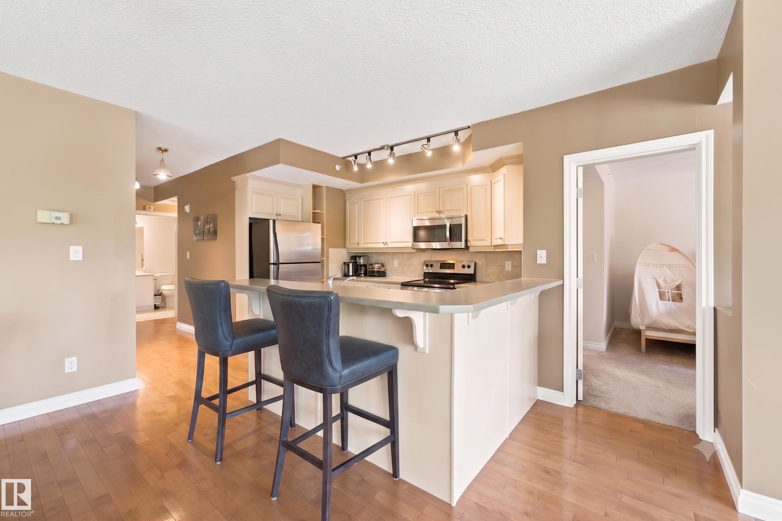 Open-concept kitchen featuring light wood-finish flooring, cream-colored cabinetry, stainless steel appliances, a breakfast bar, and track lighting - 305 11111 82 Avenue, Edmonton, AB