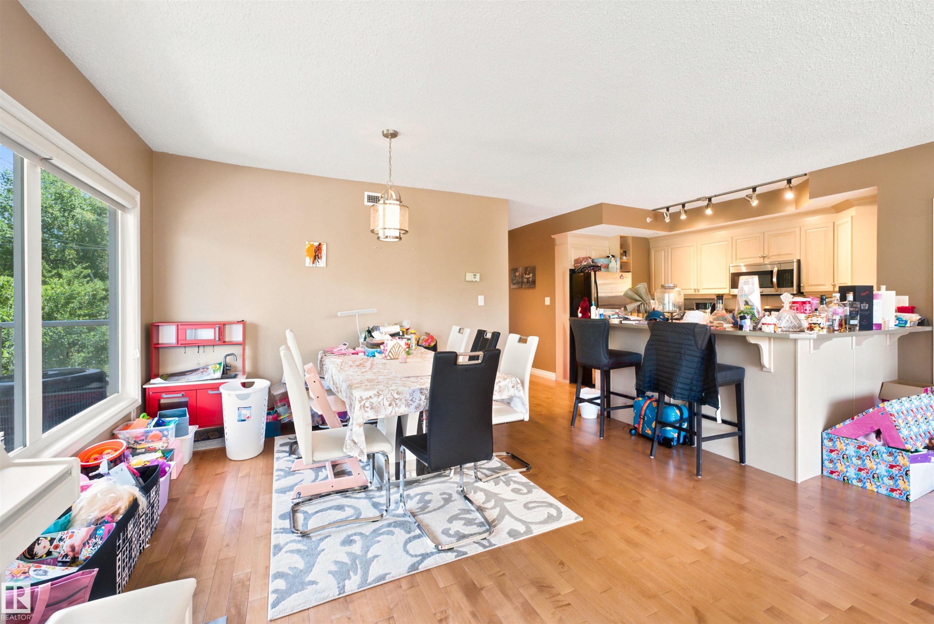 Open-concept living space featuring wood-finish flooring, a large window, and a kitchen with light cabinetry, stainless steel appliances, and a breakfast bar with seating - 305 11111 82 Avenue, Edmonton, AB