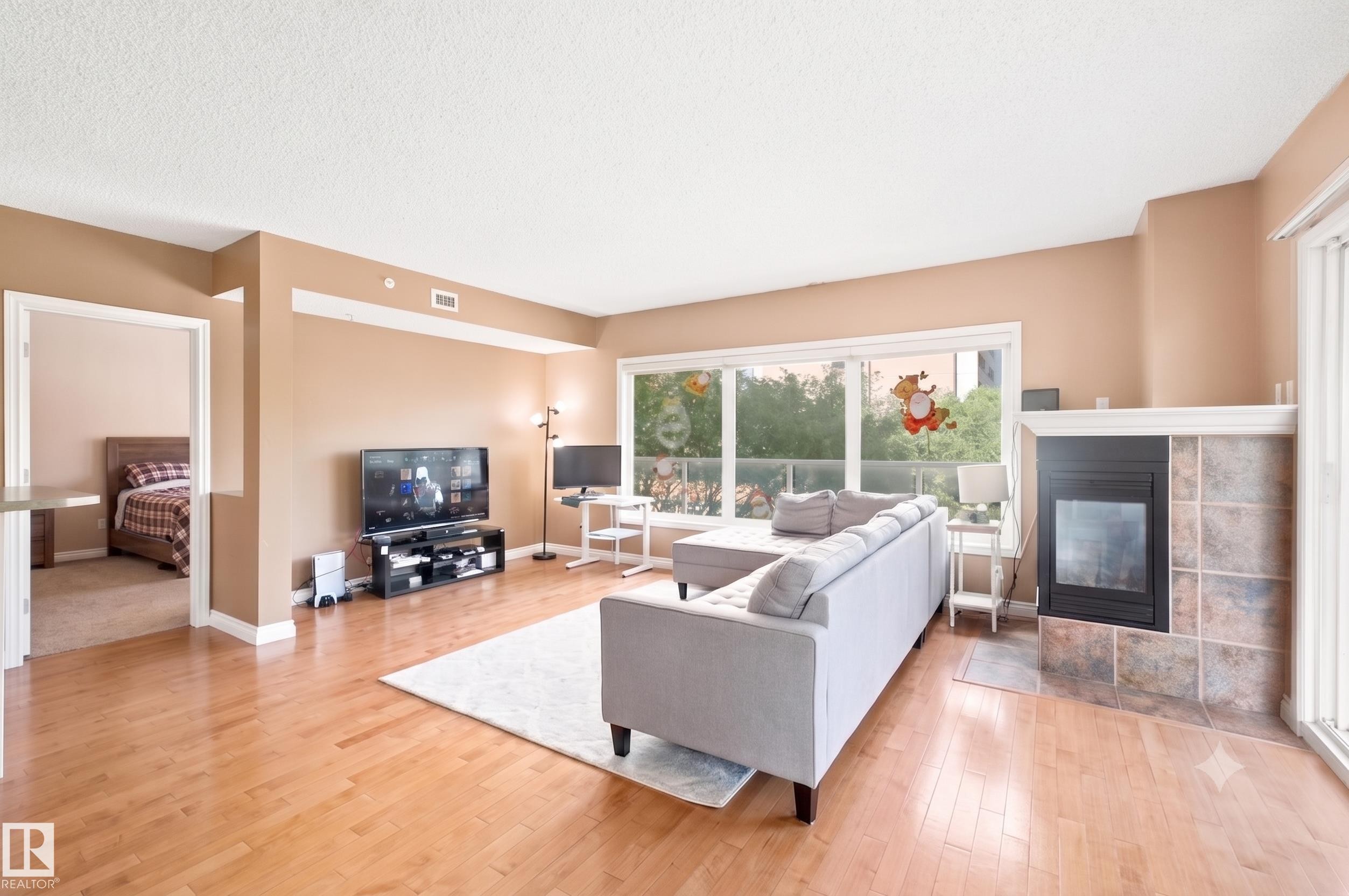 Open-concept living area featuring wood-finish flooring, a tiled fireplace with white mantle, and expansive windows - 305 11111 82 Avenue, Edmonton, AB