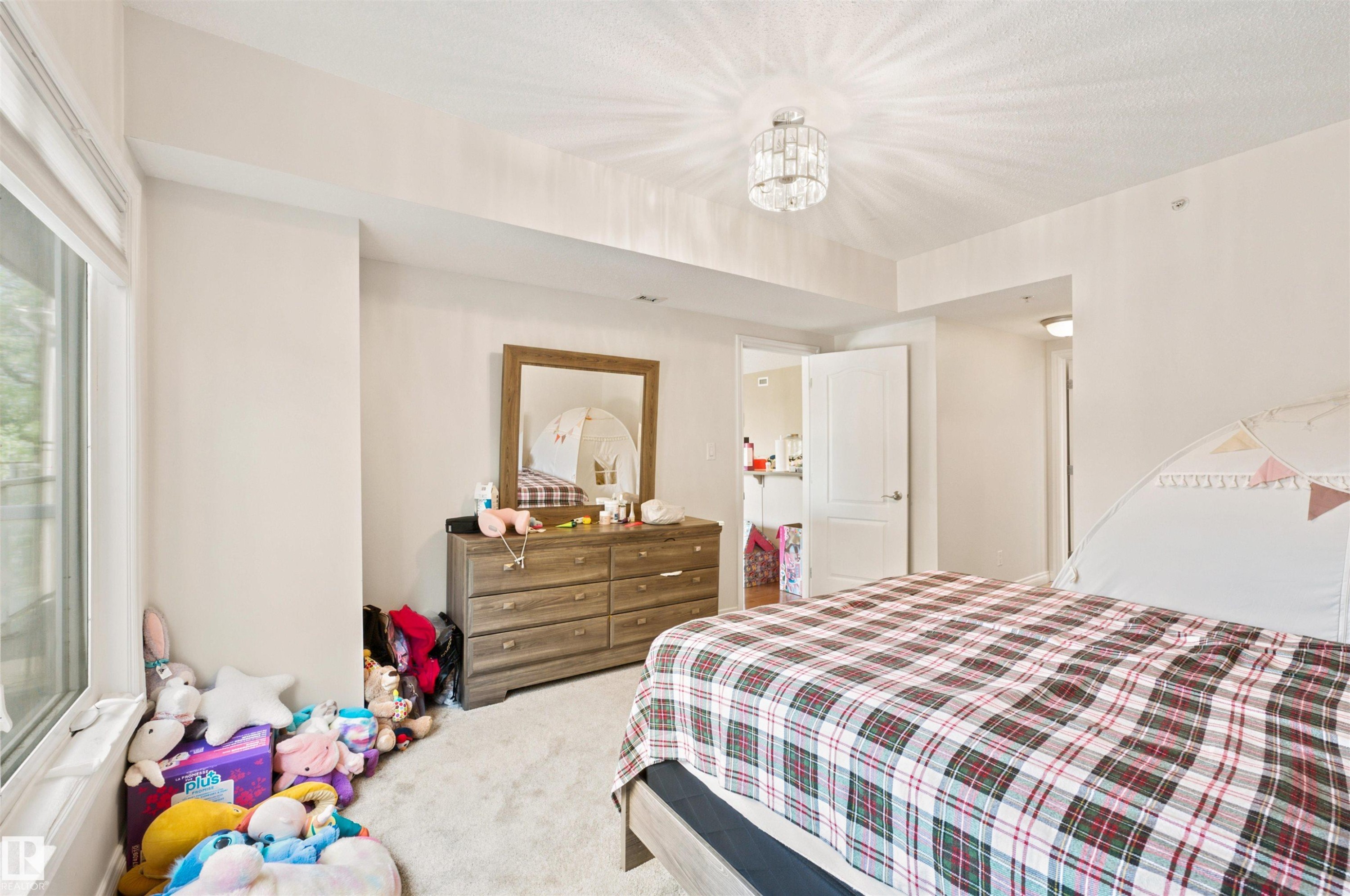 Spacious bedroom featuring light-colored carpeting, a large window with white trim, and a contemporary ceiling light fixture - 305 11111 82 Avenue, Edmonton, AB
