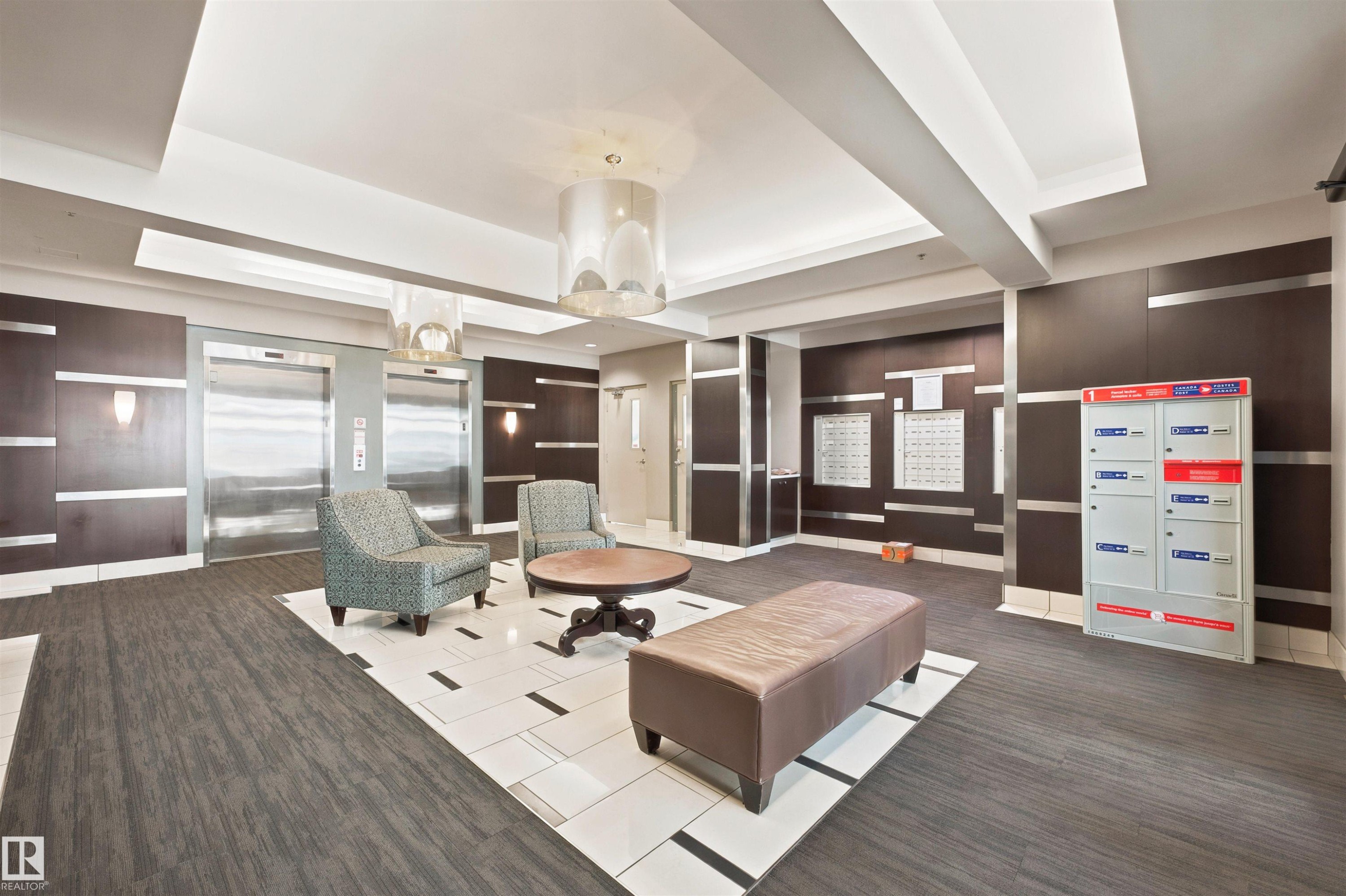 Lobby area featuring two stainless steel elevators, overhead lighting, dark wood-finish wall panels with metallic accents, and a central seating arrangement - 305 11111 82 Avenue, Edmonton, AB