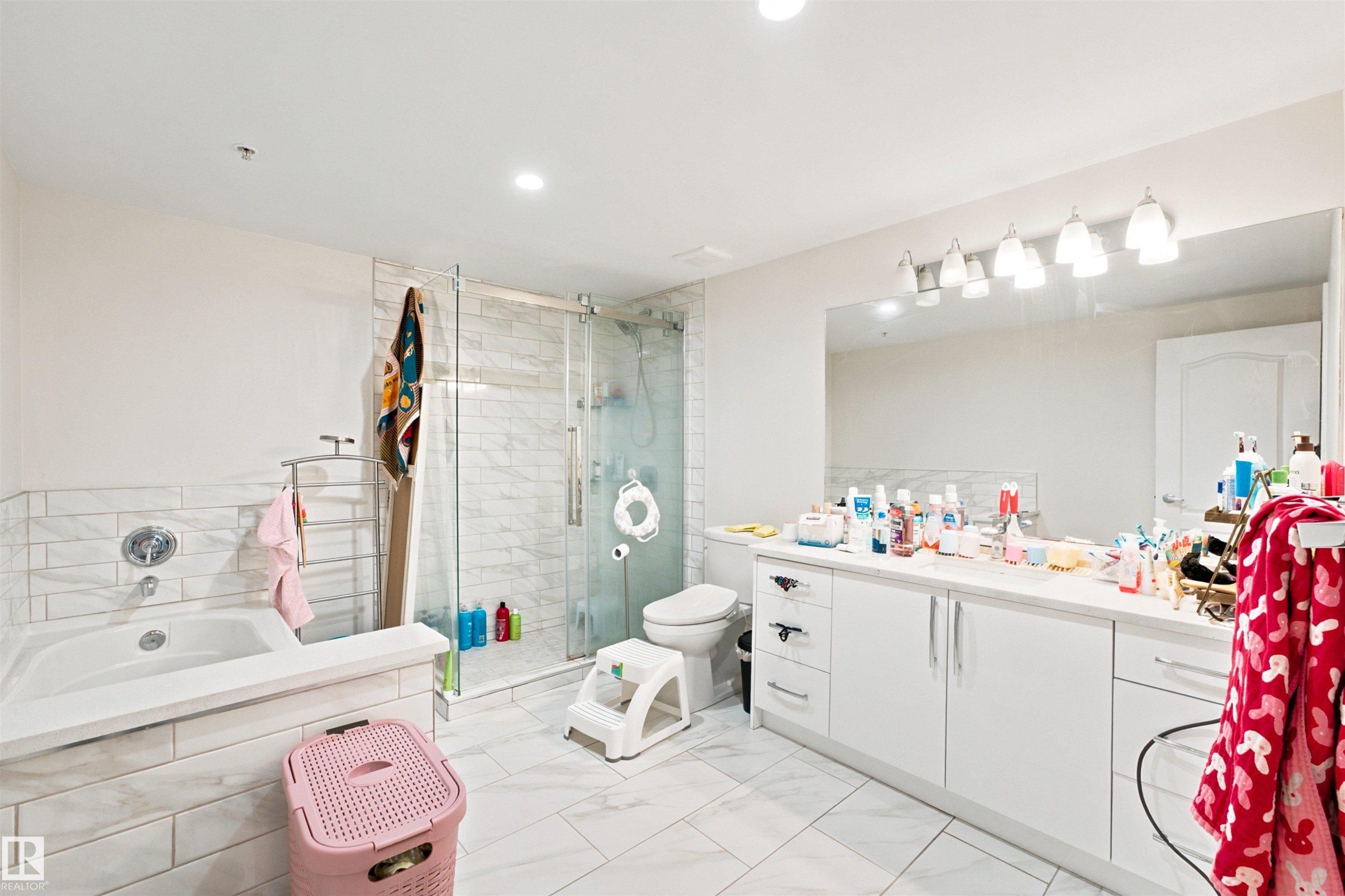 Spacious bathroom featuring a white vanity with extensive counter space, an integrated tub, a separate glass-enclosed shower, white tile flooring, and recessed lighting - 305 11111 82 Avenue, Edmonton, AB