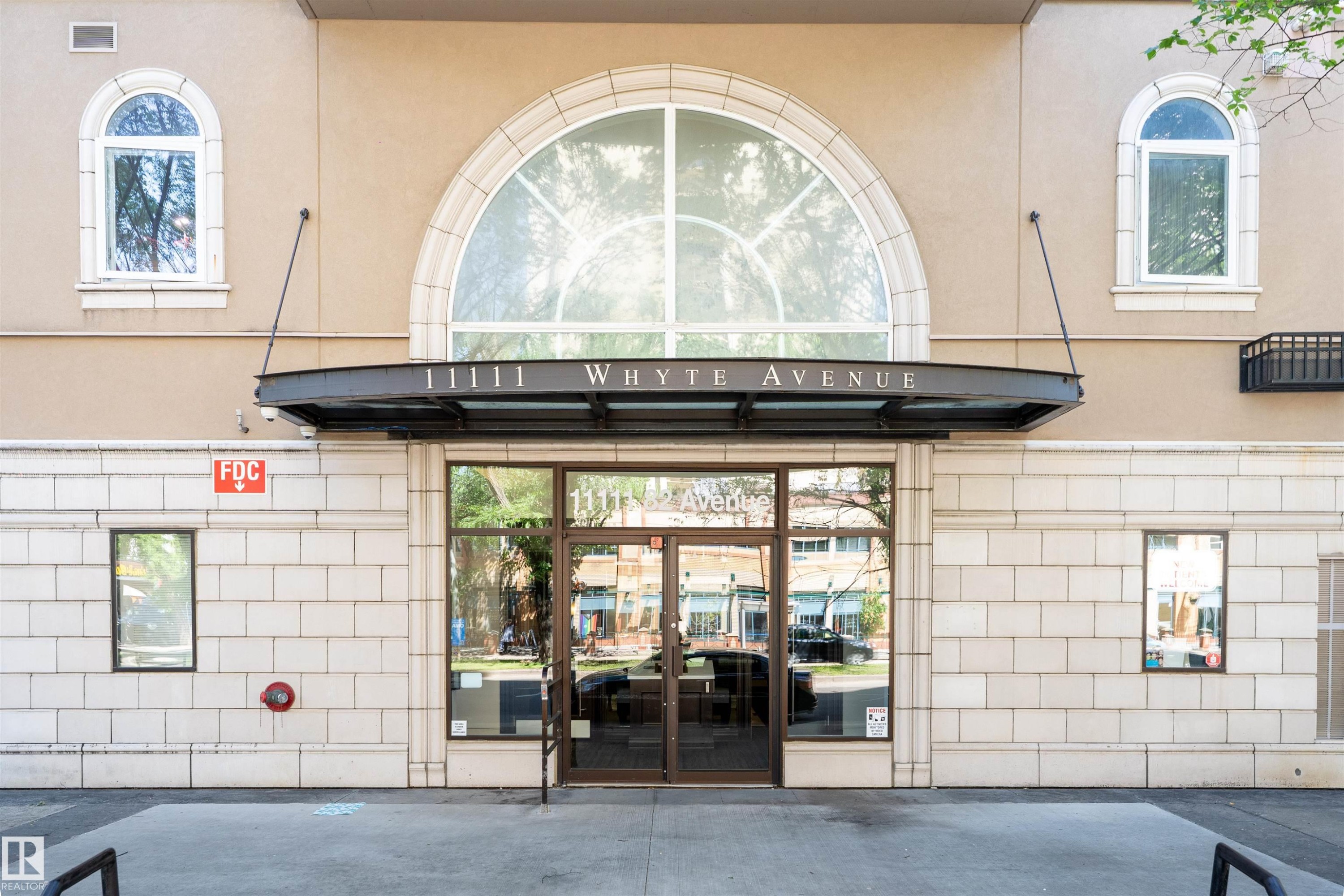 Modern building entrance featuring a prominent arched window, glass double doors, a metal canopy, and a stone facade - 305 11111 82 Avenue, Edmonton, AB